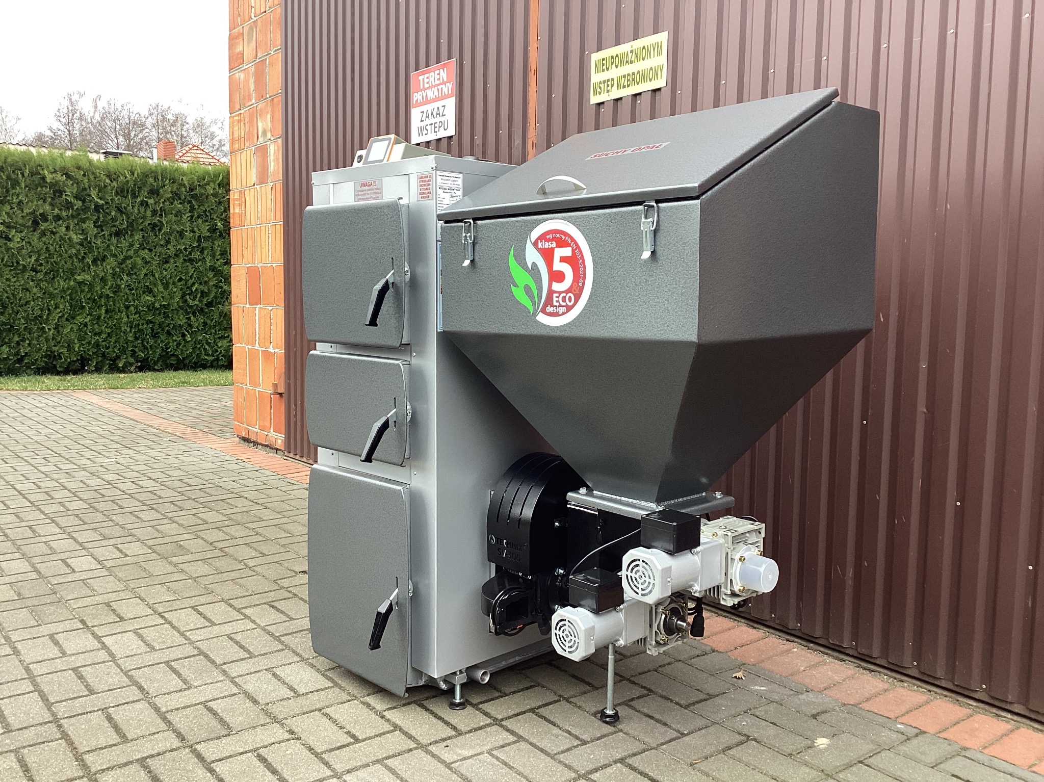 Szary piec na paliwo stałe z podajnikiem, z oznaczeniem klasy 5 Eco Design, ustawiony na kostce brukowej przed brązową bramą garażową.