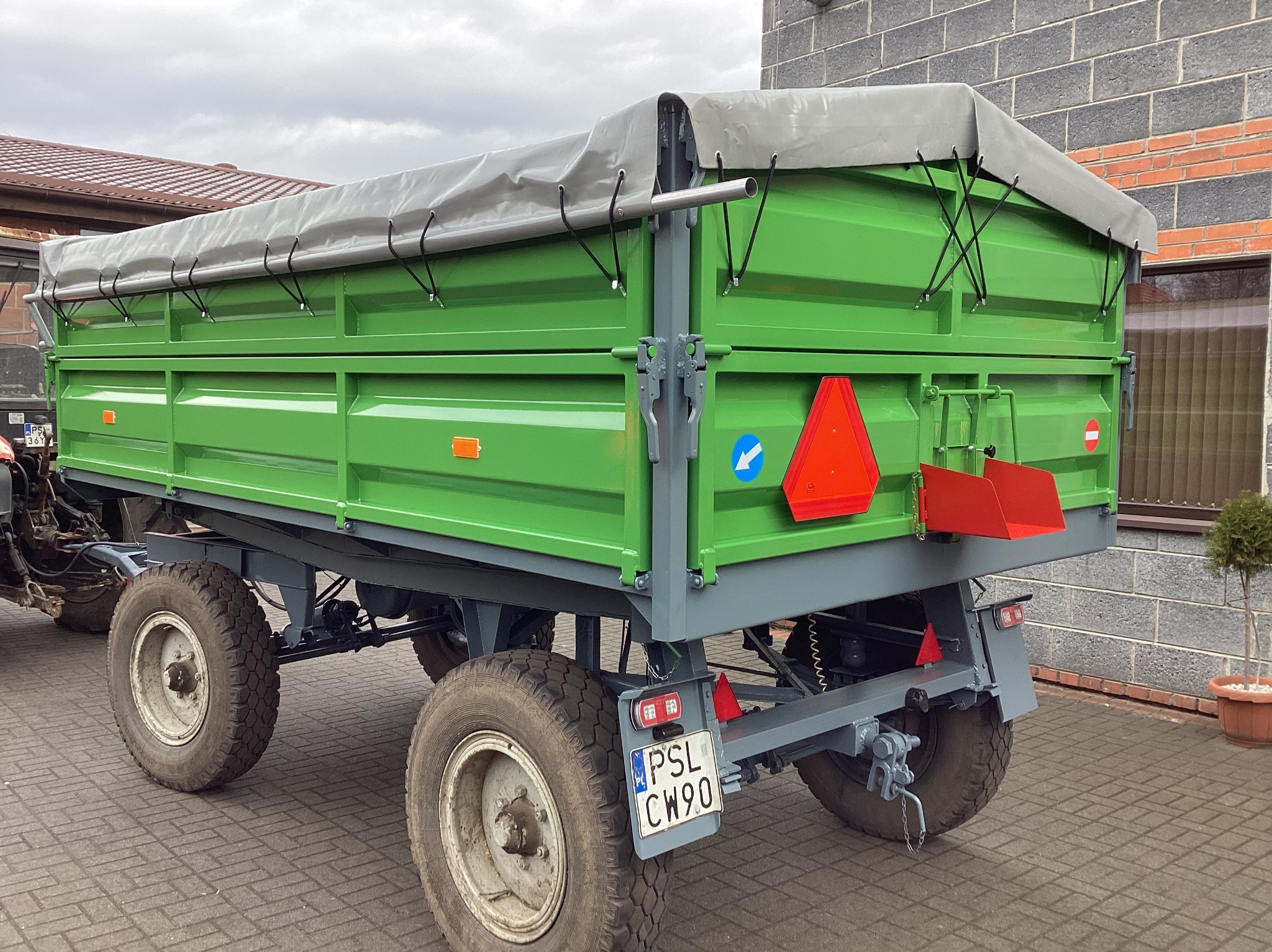 Zielona przyczepa rolnicza z szarą plandeką, widoczna tablica rejestracyjna PSL CW90, na tle brukowanej nawierzchni i ściany z szarej cegły.