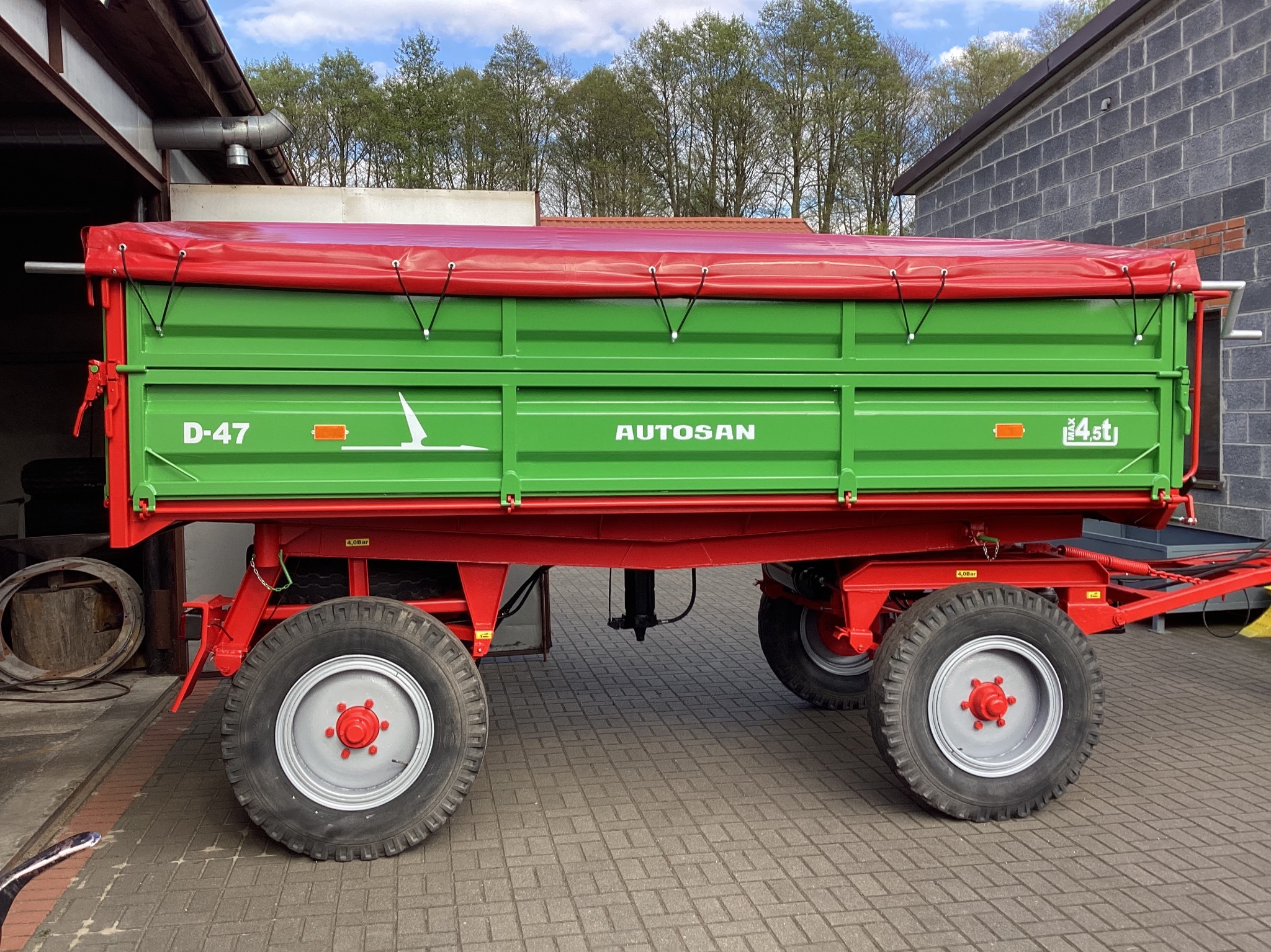 Zielono-czerwona przyczepa rolnicza Autosan D-47 z plandeką, widok z boku na tle budynku i drzew. Solidna konstrukcja, opony z szarymi felgami i czerwonymi nakrętkami.