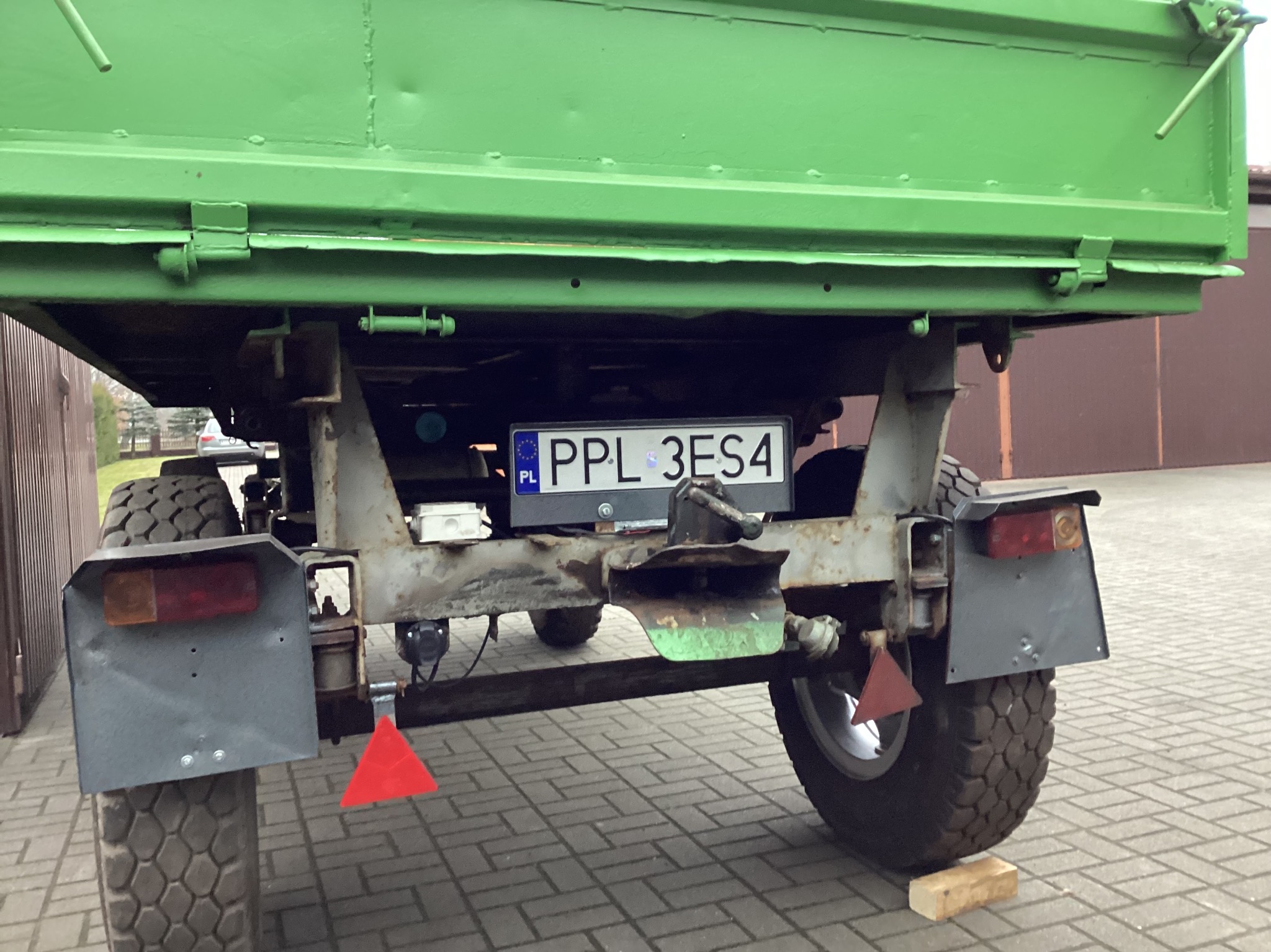 Zielona przyczepa rolnicza z widocznym numerem rejestracyjnym PPL 3ES4, opony z błotnikami i elementami konstrukcji stalowej. Widok z tyłu na kostce brukowej.