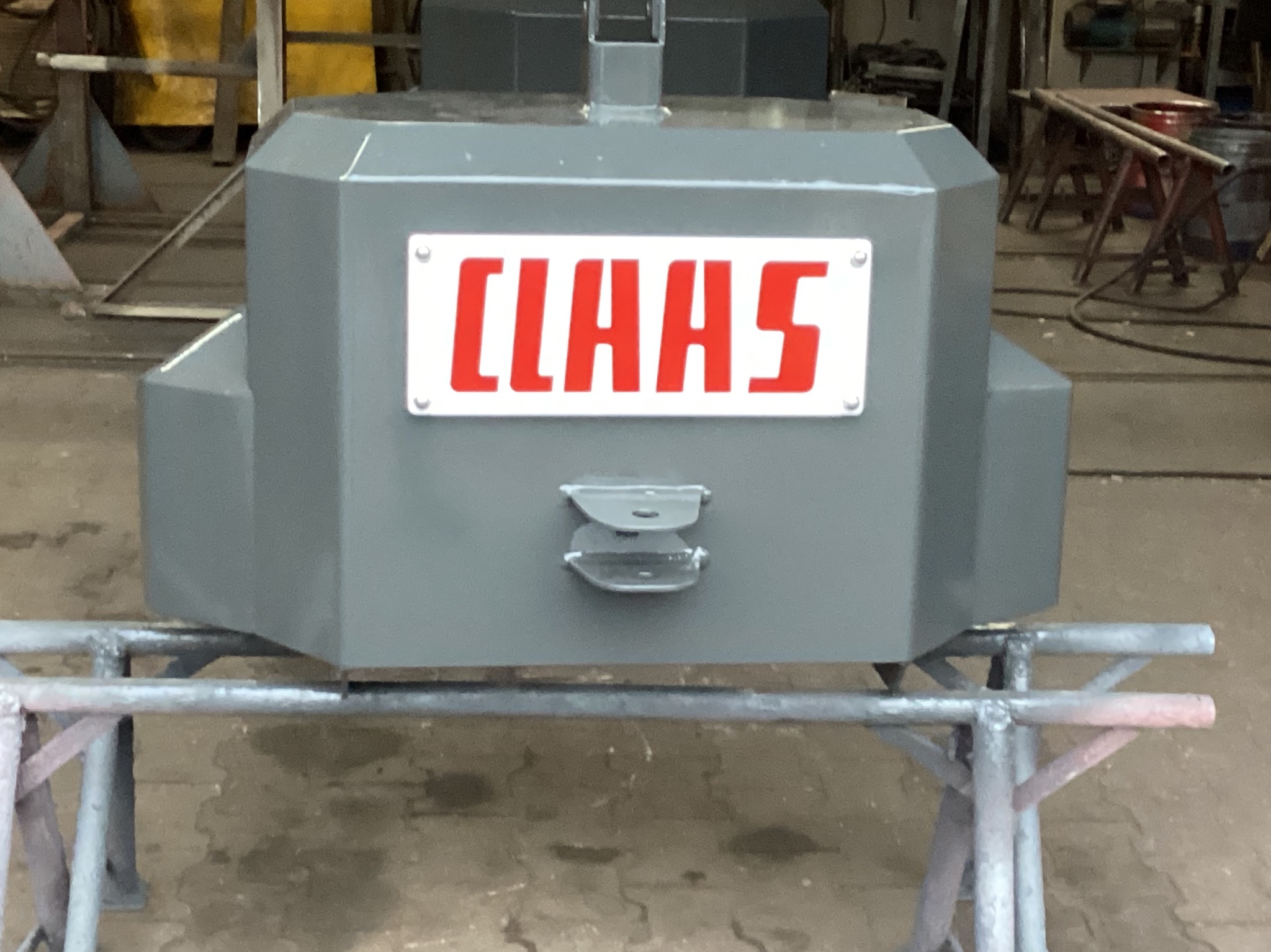 Szara, metalowa konstrukcja z napisem CLAAS na białej tabliczce, umieszczona na metalowym stelażu. Widoczne ślady spawania i obróbki metalu w tle warsztatu.