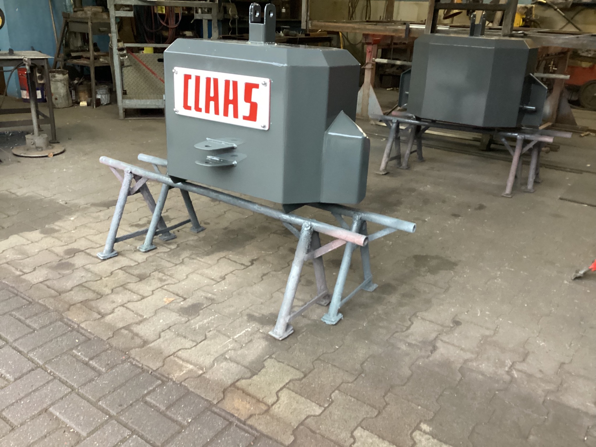 Szary, metalowy obciążnik z logo CLAAS, umieszczony na metalowym stojaku w warsztacie. Widoczny drugi obciążnik w tle. Podłoże z kostki brukowej.