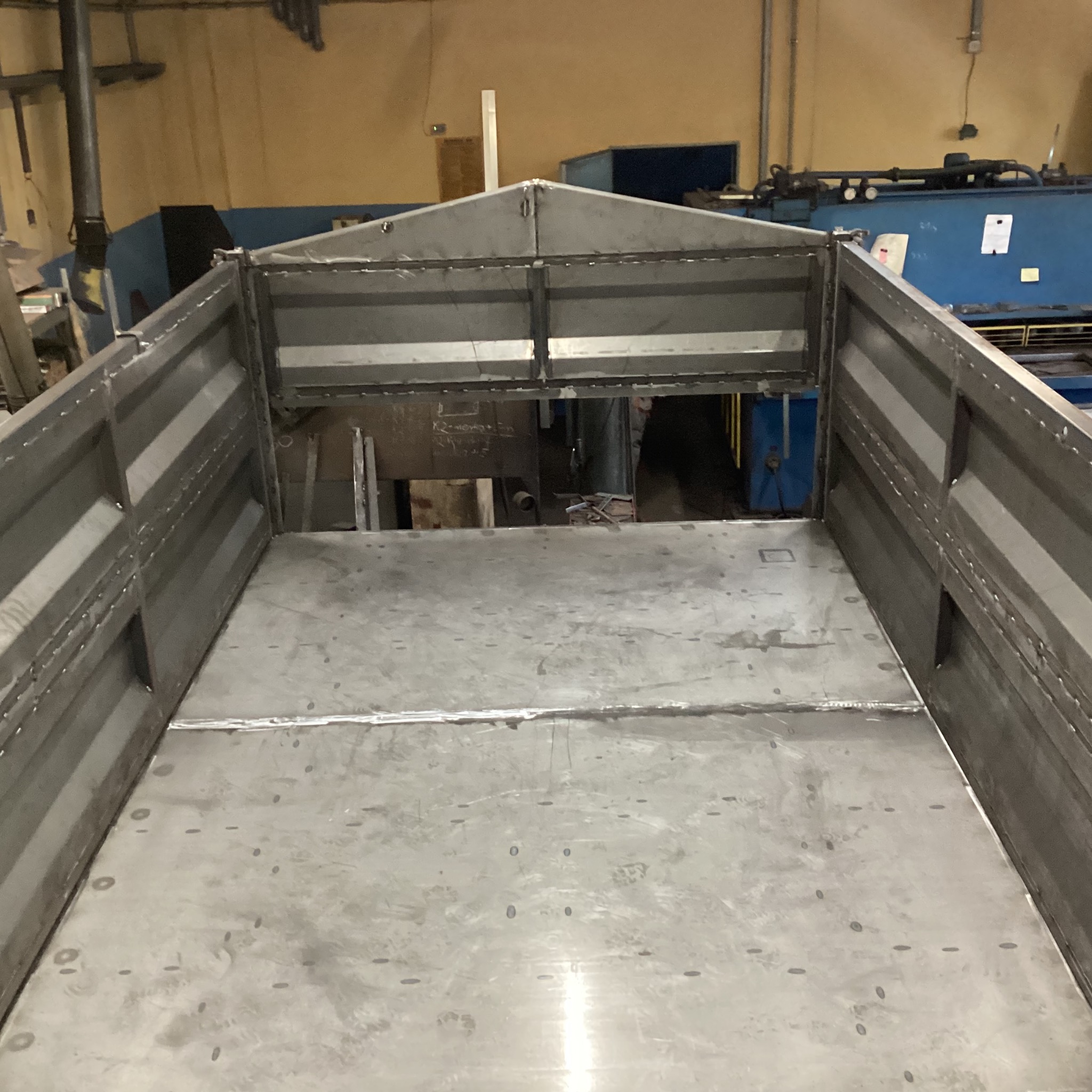 Wnętrze metalowej konstrukcji skrzyniowej z widocznymi spawami i nitami, wykonanej w warsztacie. Widok z góry na surową, metalową powierzchnię.