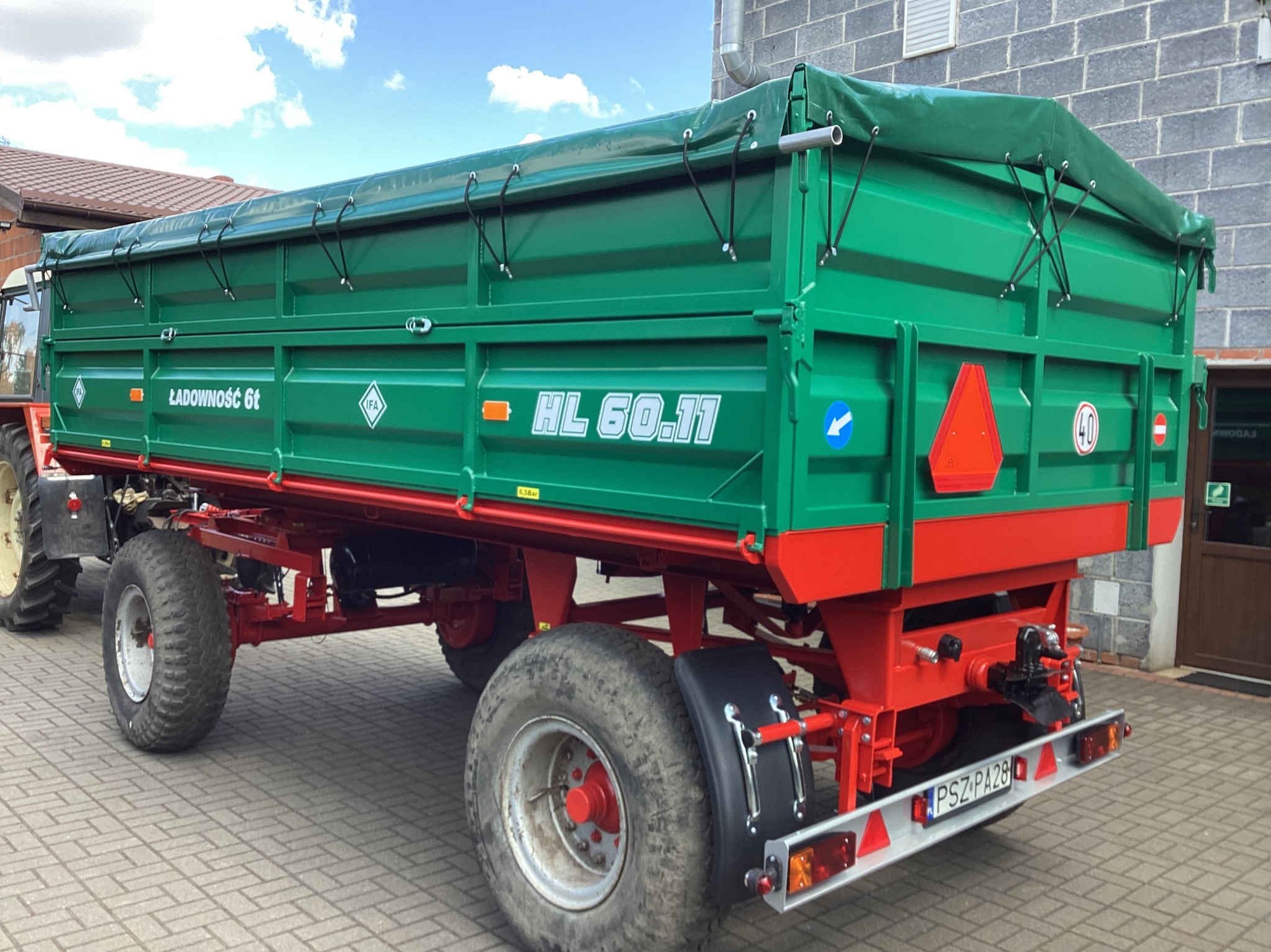 Zielono-czerwona przyczepa rolnicza HL 60.11 z plandeką, widoczna tablica rejestracyjna. Z tyłu fragment ciągnika i budynek z szarej cegły.