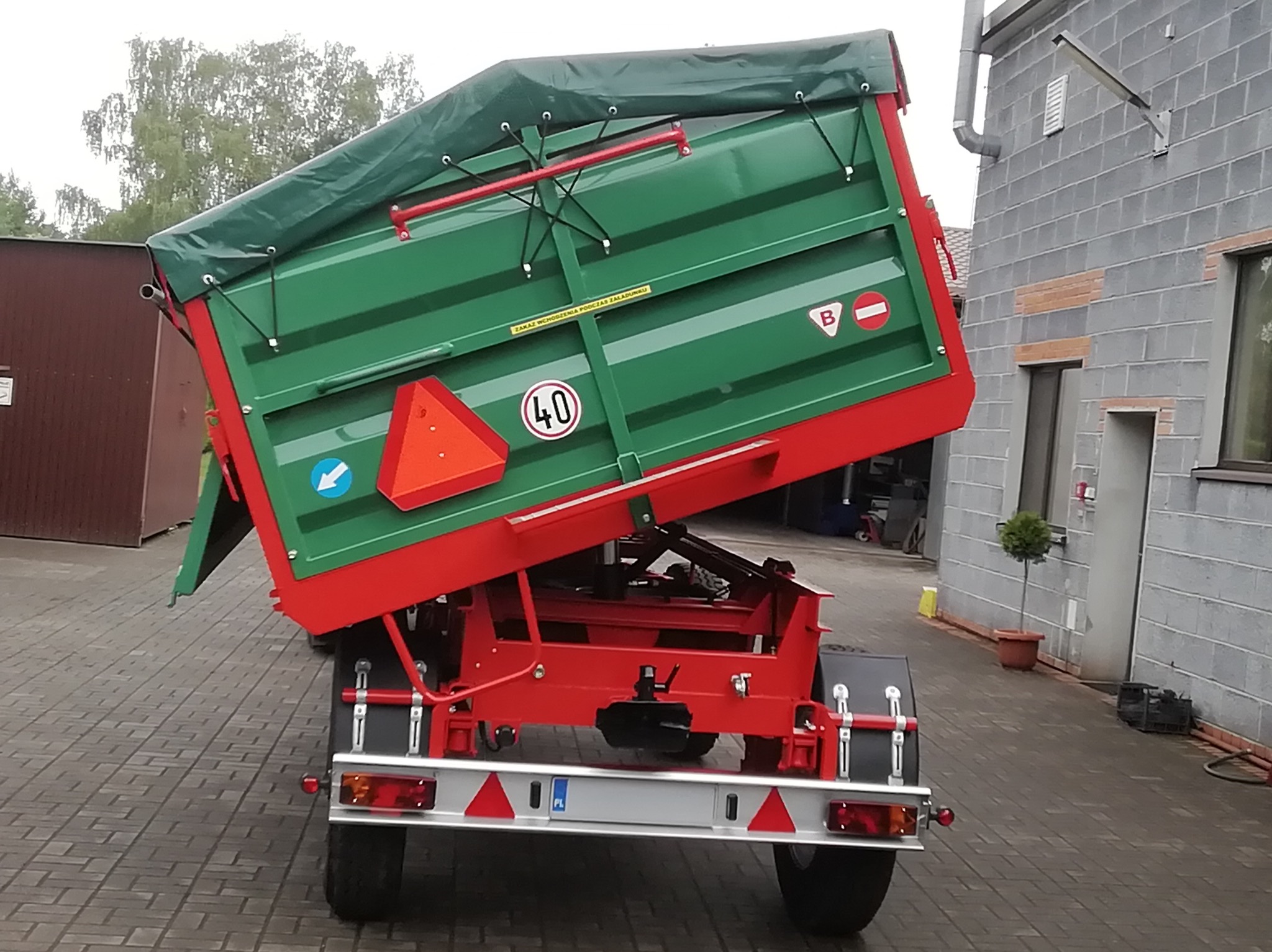 Podniesiona, zielono-czerwona przyczepa rolnicza z plandeką, widoczna z tyłu na tle szarego budynku i kostki brukowej. Tablice ostrzegawcze i ograniczenie prędkości 40 km/h.