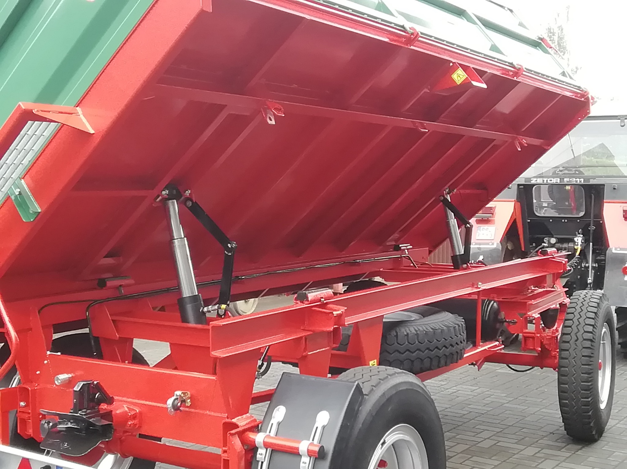 Czerwona, podniesiona przyczepa rolnicza z widocznymi siłownikami hydraulicznymi i kołami, zaparkowana na kostce brukowej. W tle widoczny ciągnik Zetor.