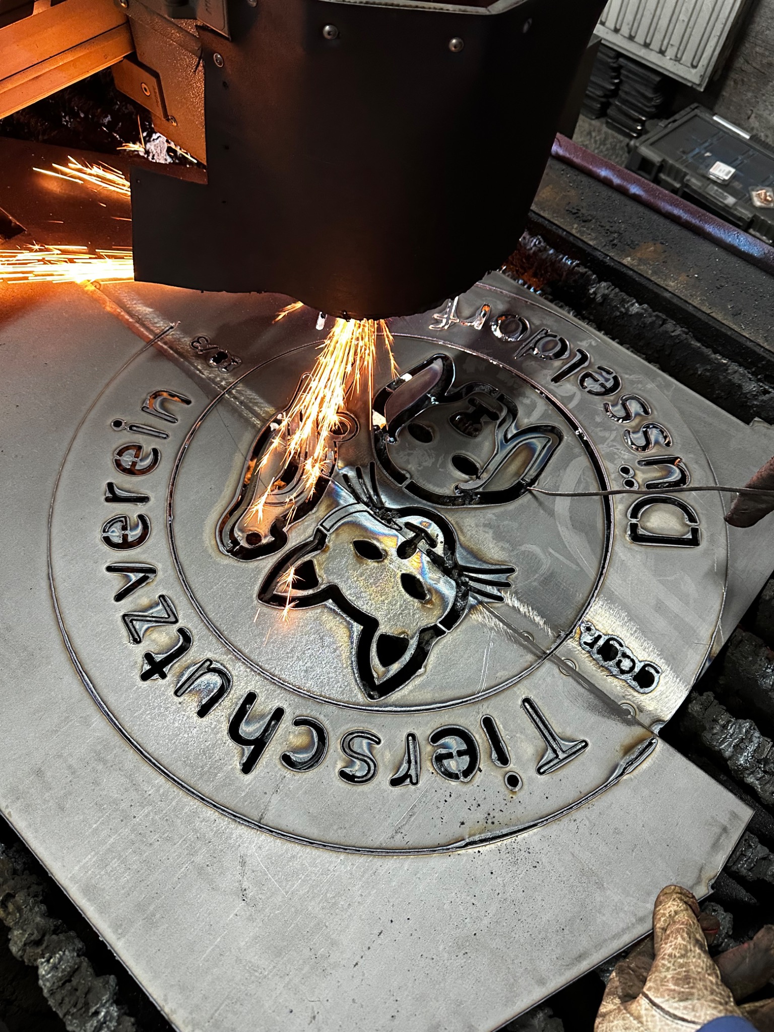 Precyzyjna obróbka CNC metalu: wycinanie plazmowe logo 'Tierschutzverein Düsseldorf' z arkusza stali, iskry, rękawica ochronna w kadrze. Detal procesu.