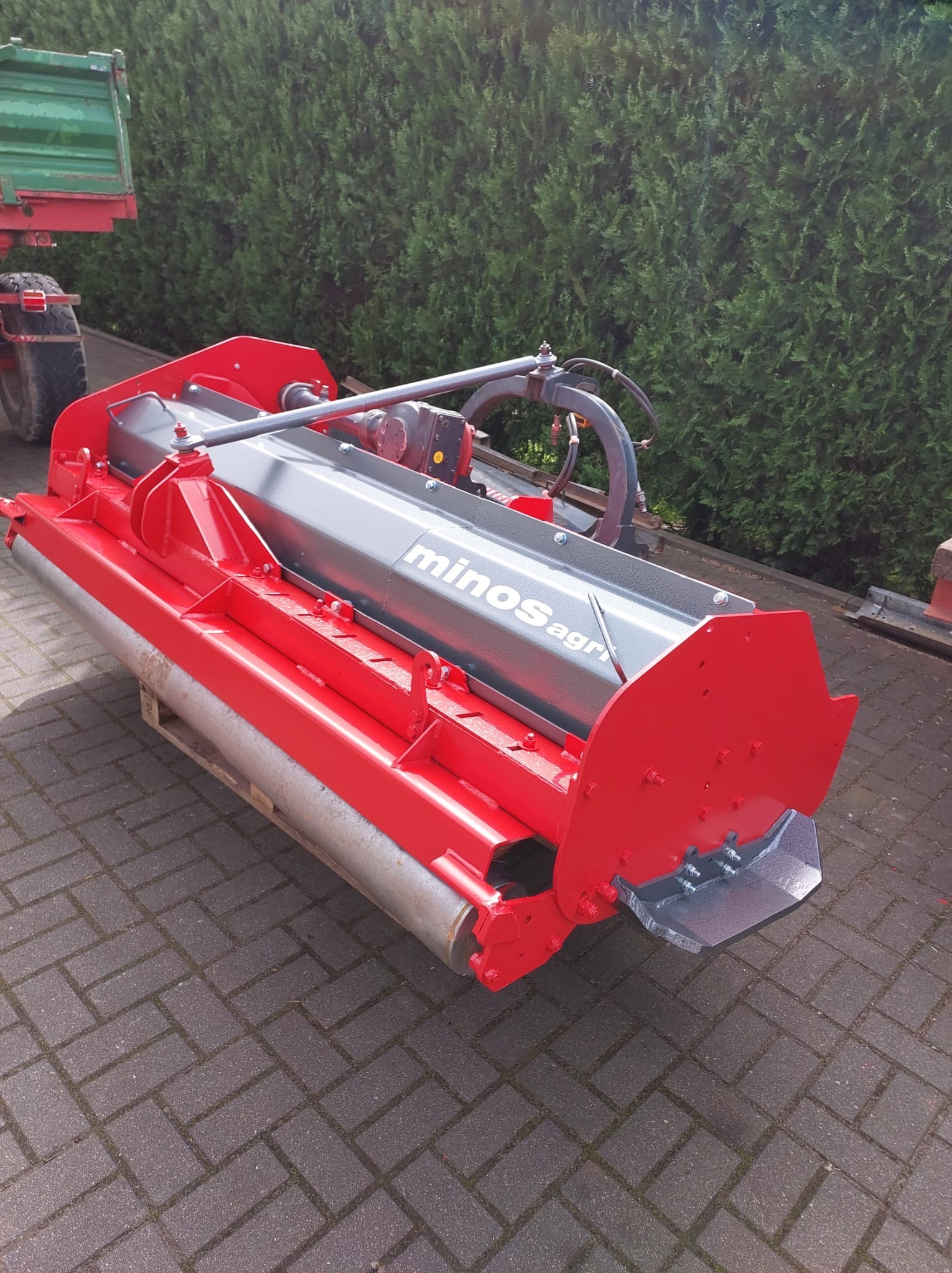 Czerwono-szara kosiarka bijakowa Minos Agri na palecie, z fragmentem traktora w tle, na kostce brukowej. Tło stanowi zielony żywopłot.