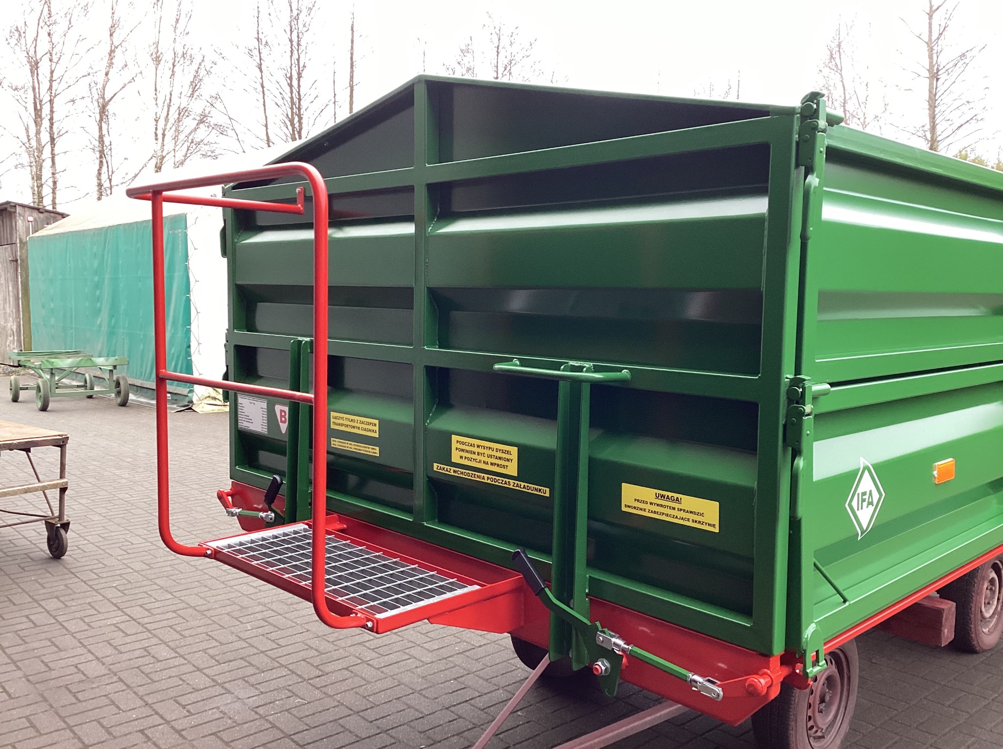 Zielona przyczepa rolnicza z czerwoną drabinką i platformą, widoczne tabliczki ostrzegawcze, na tle drzew i budynków gospodarczych. Detal konstrukcji.
