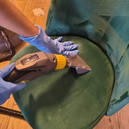 ABM Pro Łukasz Motłoch - Czyszczenie welurowego krzesła na drewnianej podłodze. Ręka w niebieskiej rękawicy obsługuje odkurzacz piorący, widoczne ślady czyszczenia na zielonym siedzisku.