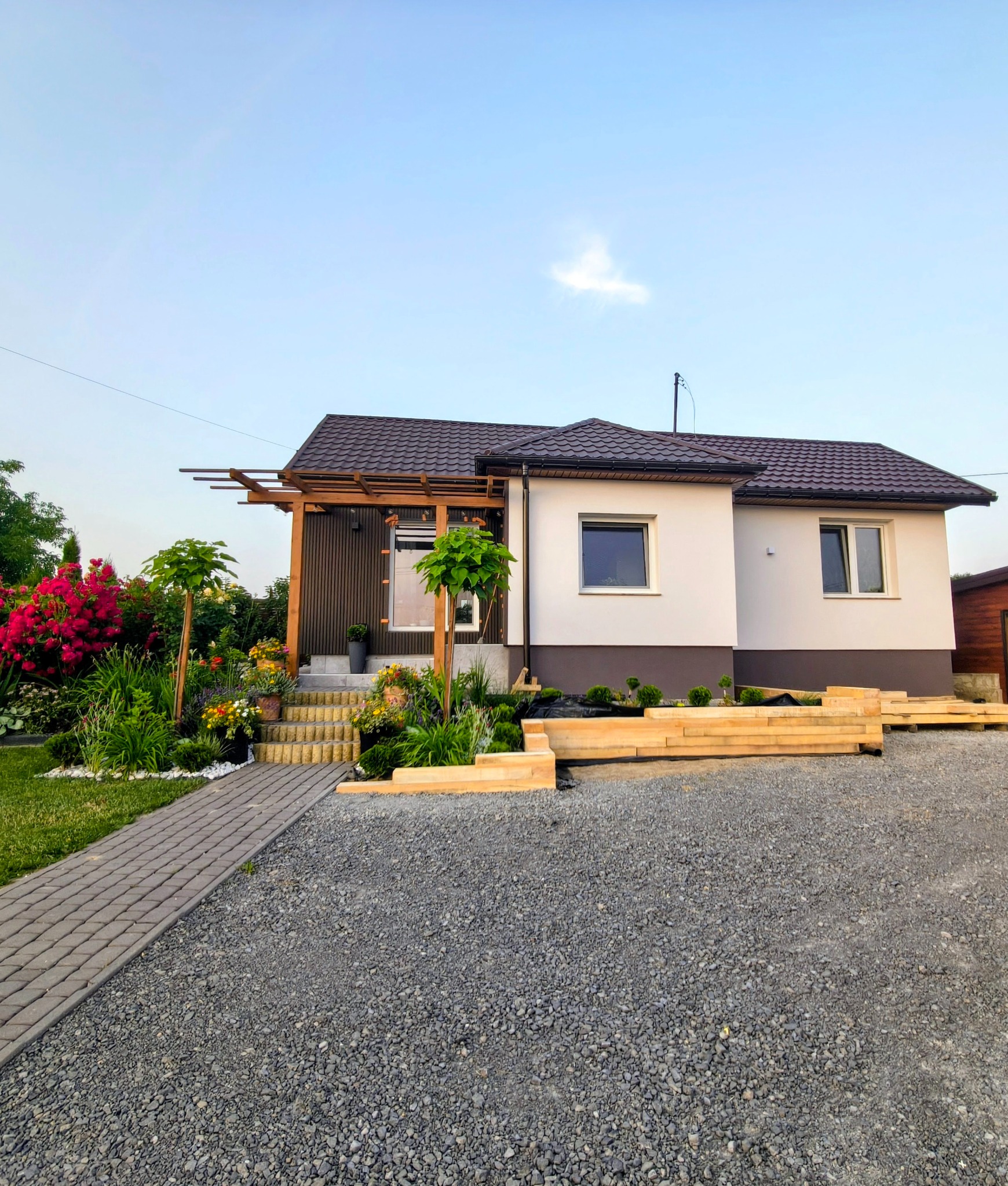 Nowoczesny dom jednorodzinny z brązowym dachem i elementami drewnianymi, otoczony zadbanym ogrodem z kostką brukową i rabatami kwiatowymi. Widok z przodu, słoneczny dzień.