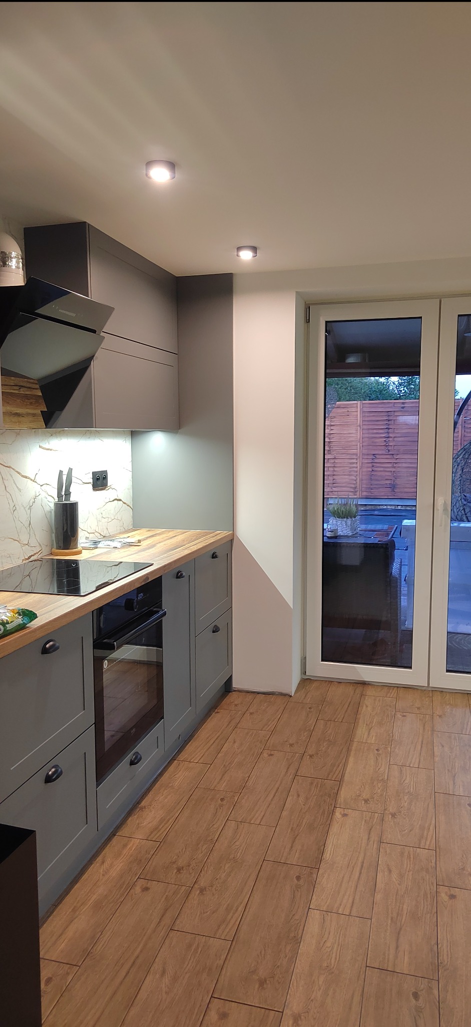 Nowoczesna kuchnia w szarościach z drewnianym blatem i podłogą. Widok na taras przez przeszklone drzwi balkonowe. Minimalistyczny design i jasne oświetlenie.