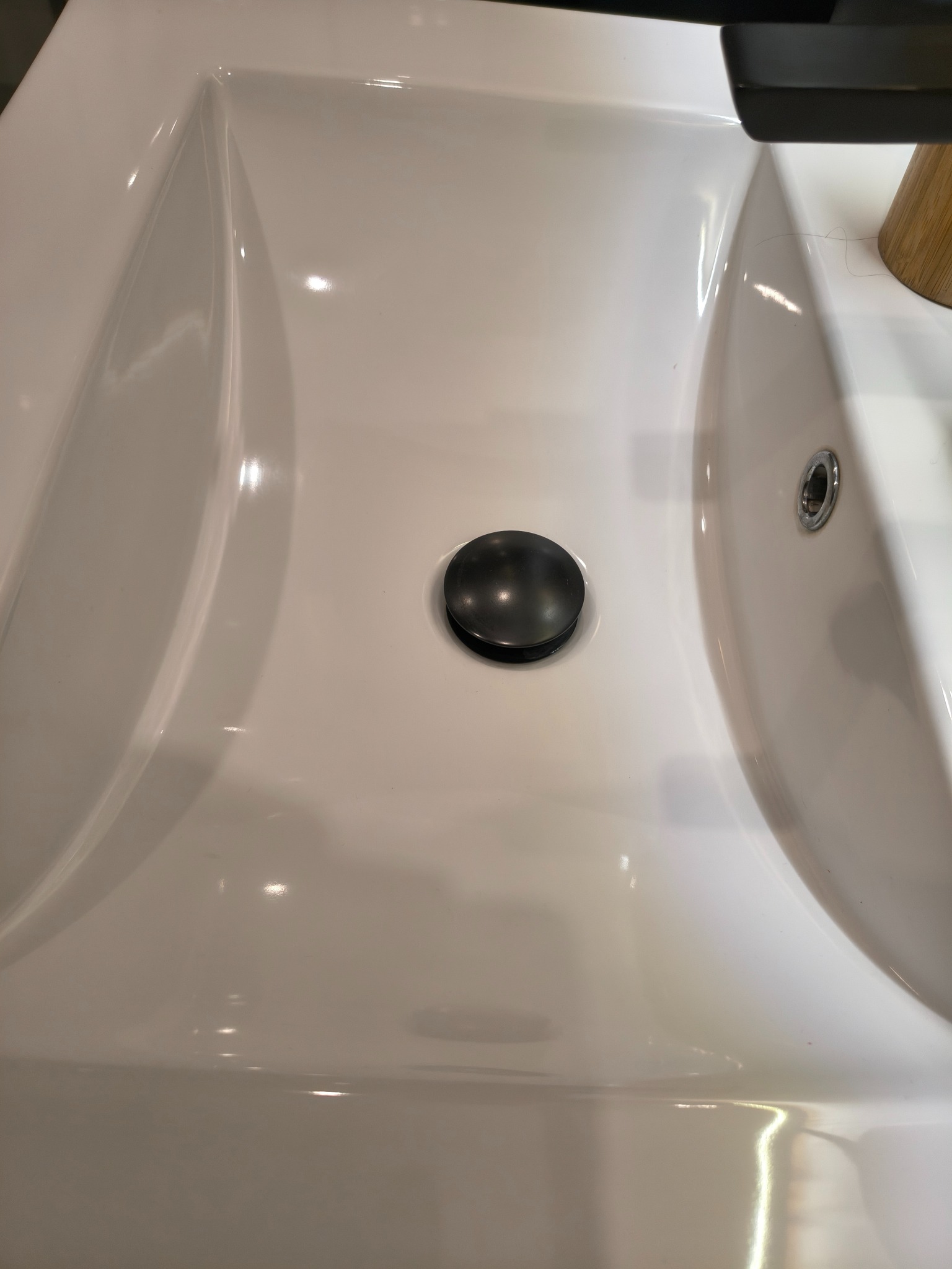 Biała, ceramiczna umywalka z czarnym korkiem spustowym i fragmentem czarnej baterii. Widoczne refleksy światła na gładkiej powierzchni.