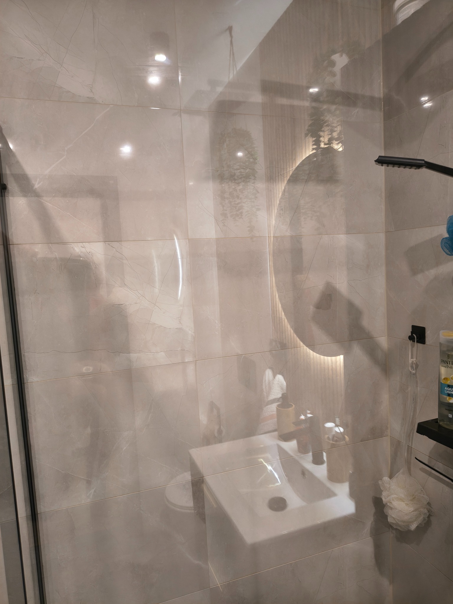 Nowoczesna łazienka z marmurowymi płytkami, podświetlanym lustrem i minimalistyczną umywalką. Widoczny fragment kabiny prysznicowej i akcesoria łazienkowe.