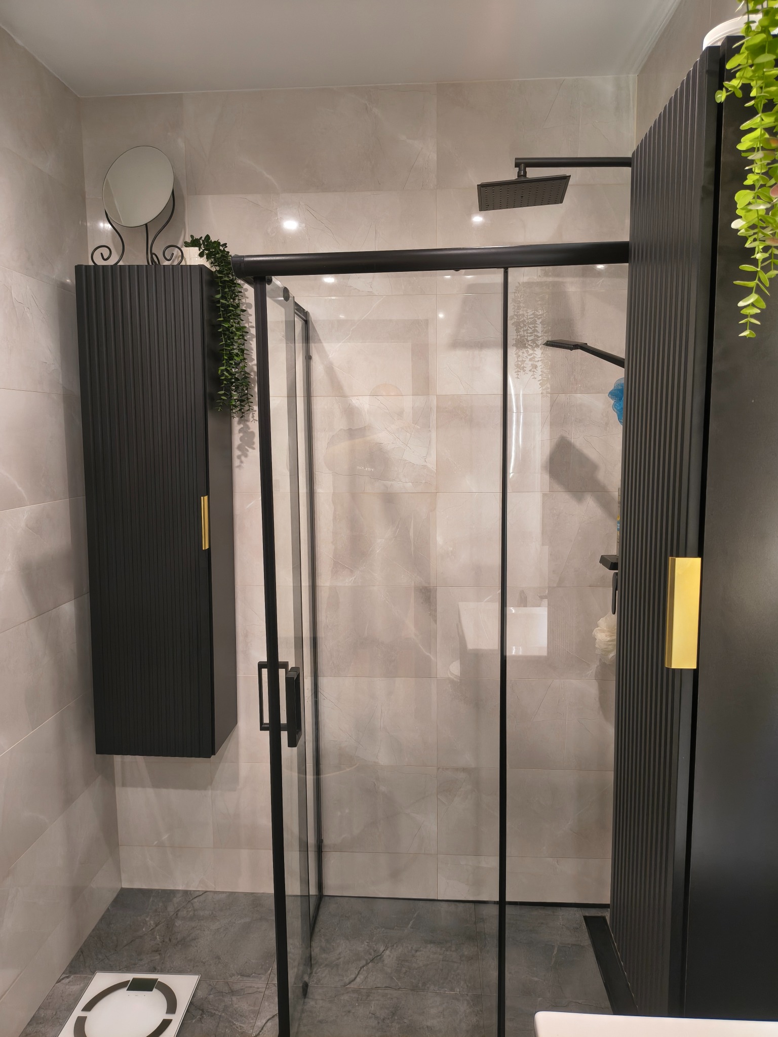 Nowoczesna kabina prysznicowa z czarnymi elementami i złotymi uchwytami, uzupełniona o wiszącą szafkę i rośliny, na tle marmurowych płytek.