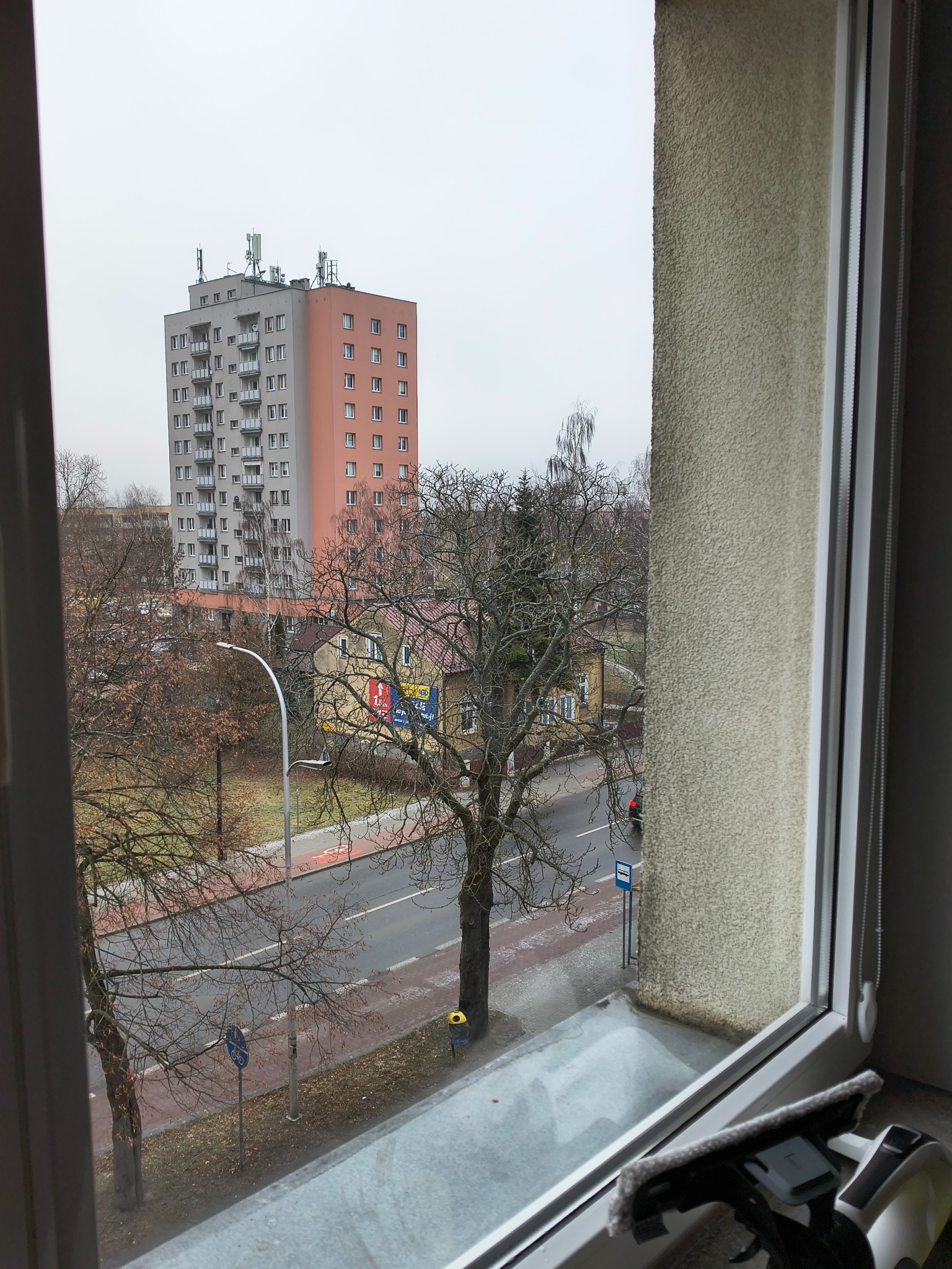 Widok z okna na blok i ulicę w pochmurny dzień. Na parapecie leży myjka do okien, sugerująca świeżo umyte okno. Perspektywa z wnętrza mieszkania.