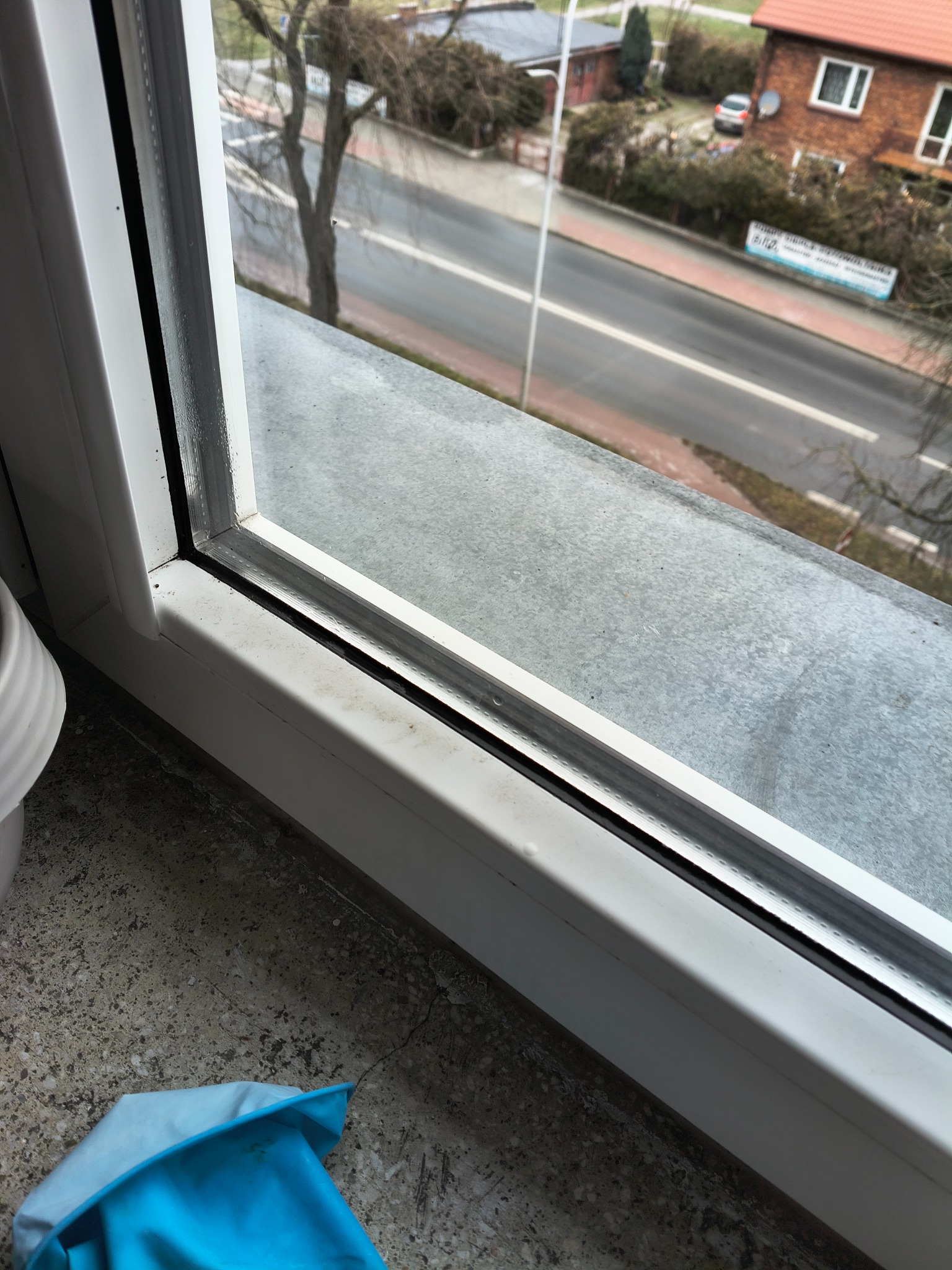 Brudne okno PCV z widocznymi smugami i zabrudzeniami na parapecie, z fragmentem niebieskiej rękawicy w rogu. Widok na ulicę zza okna.