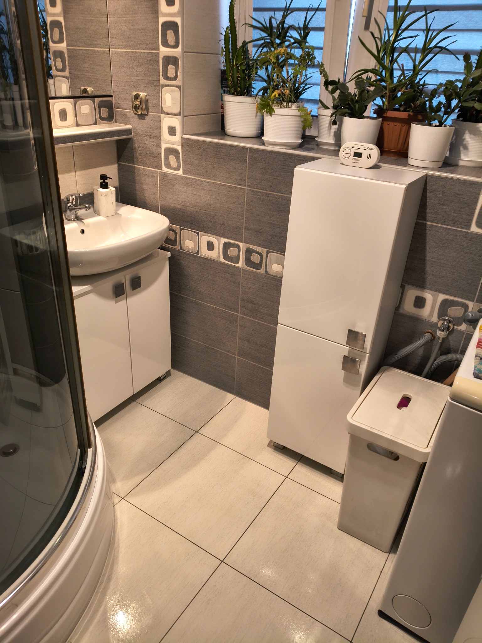 Jasna łazienka po remoncie: białe meble, umywalka z szafką, kabina prysznicowa, rośliny w doniczkach na parapecie. Czyste płytki podłogowe i ścienne.