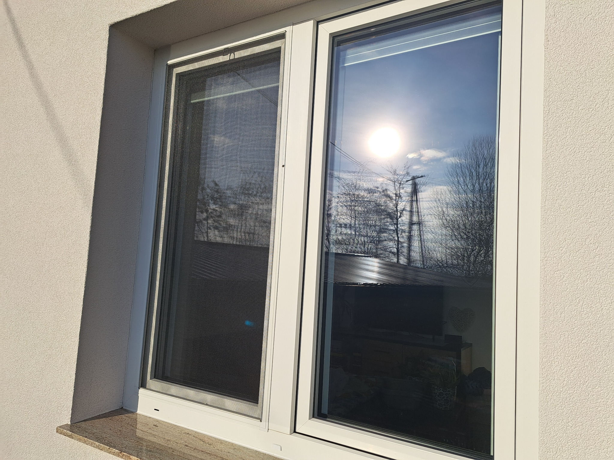 Białe, dwuskrzydłowe okno z moskitierą po lewej stronie, odbijające słońce i drzewa. Widoczny fragment elewacji budynku o jasnej, ziarnistej strukturze.