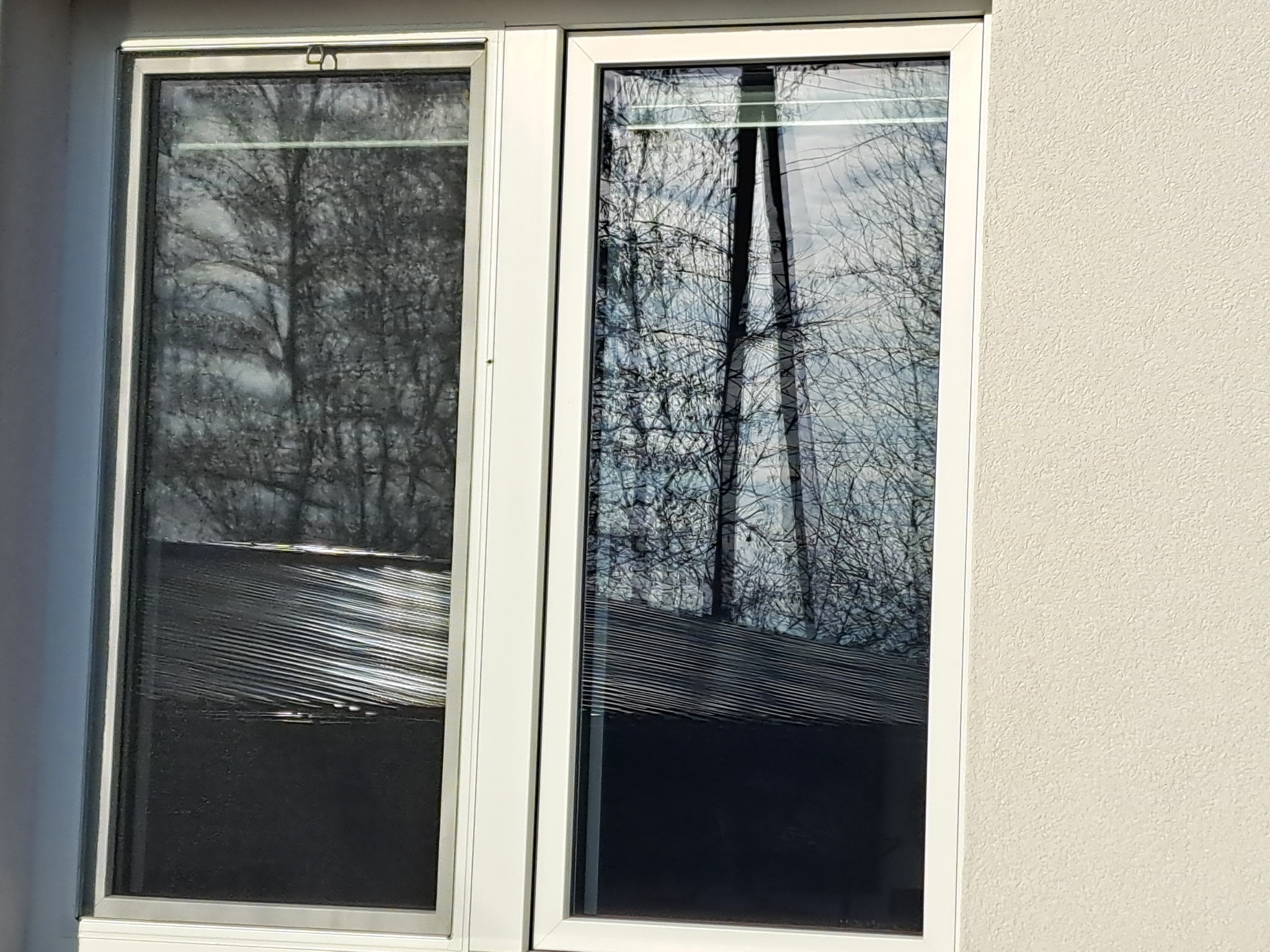 Dwuokienne okno PCV. Jedna szyba brudna, z widocznymi smugami i zaciekami, druga czysta, odbija drzewa. Biała rama okienna na tle jasnej, teksturowanej ściany.