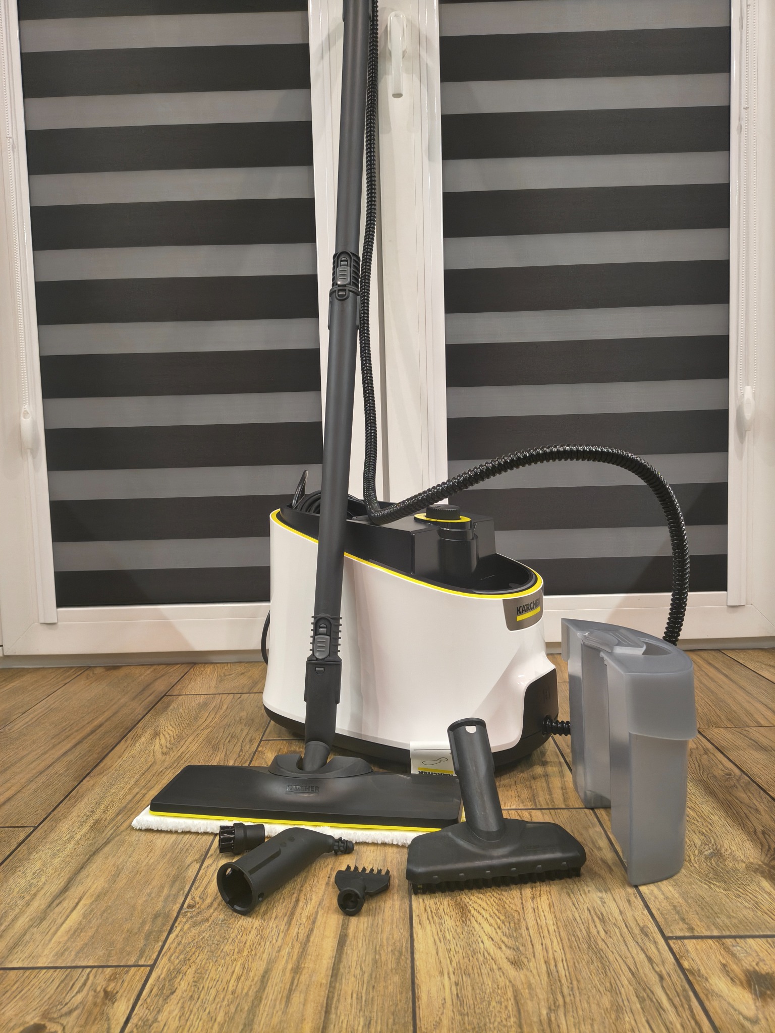 Parownica Kärcher z akcesoriami na drewnianej podłodze, na tle okna z żaluzjami. Prezentacja sprzętu do czyszczenia parowego.