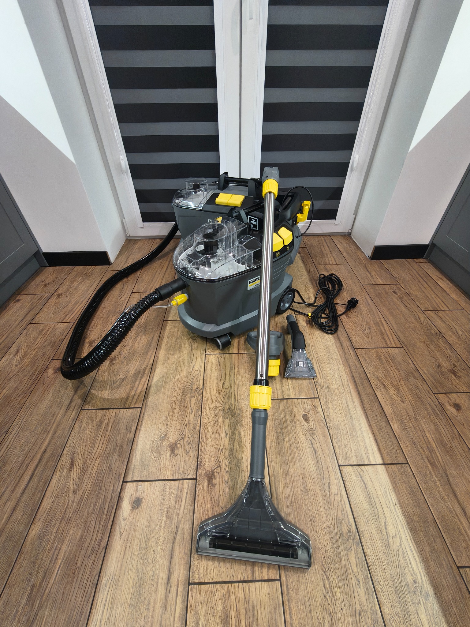 Profesjonalny odkurzacz piorący Karcher z akcesoriami na drewnianej podłodze, gotowy do prania dywanów lub tapicerki. Widoczne okno i rolety w tle.