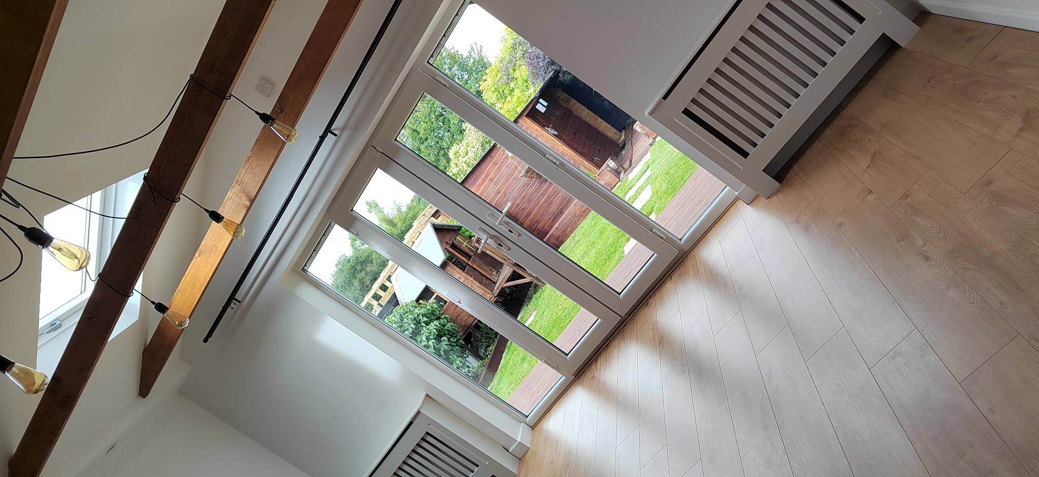 Jasne wnętrze z drewnianą podłogą i dużymi oknami wychodzącymi na zielony ogród. Widoczne belki stropowe z żarówkami i grzejnik pod oknem.