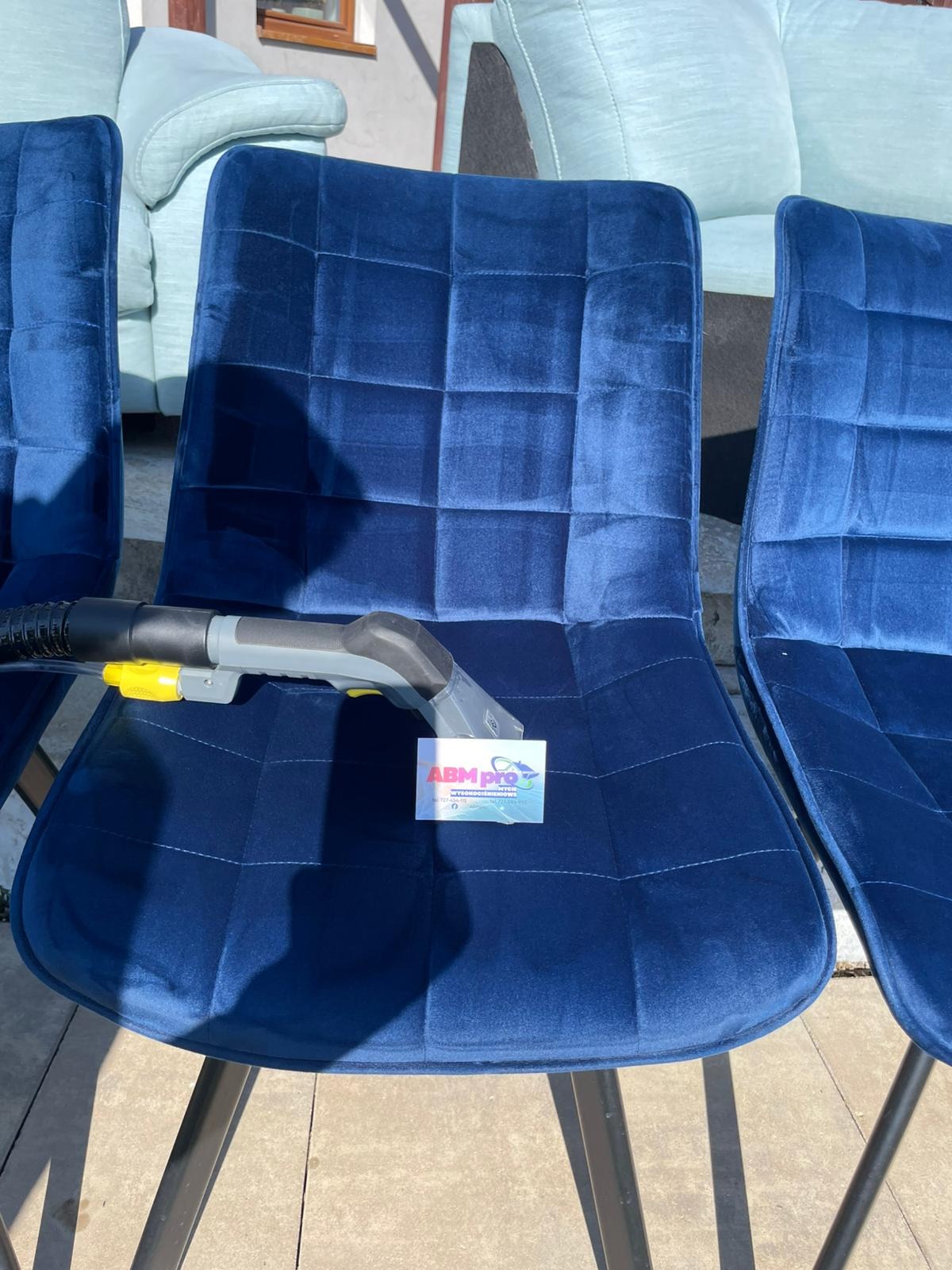 Pranie ekstrakcyjne granatowego krzesła z welurową tapicerką. Widoczna dysza odkurzacza piorącego i wizytówka firmy. W tle jasna sofa.