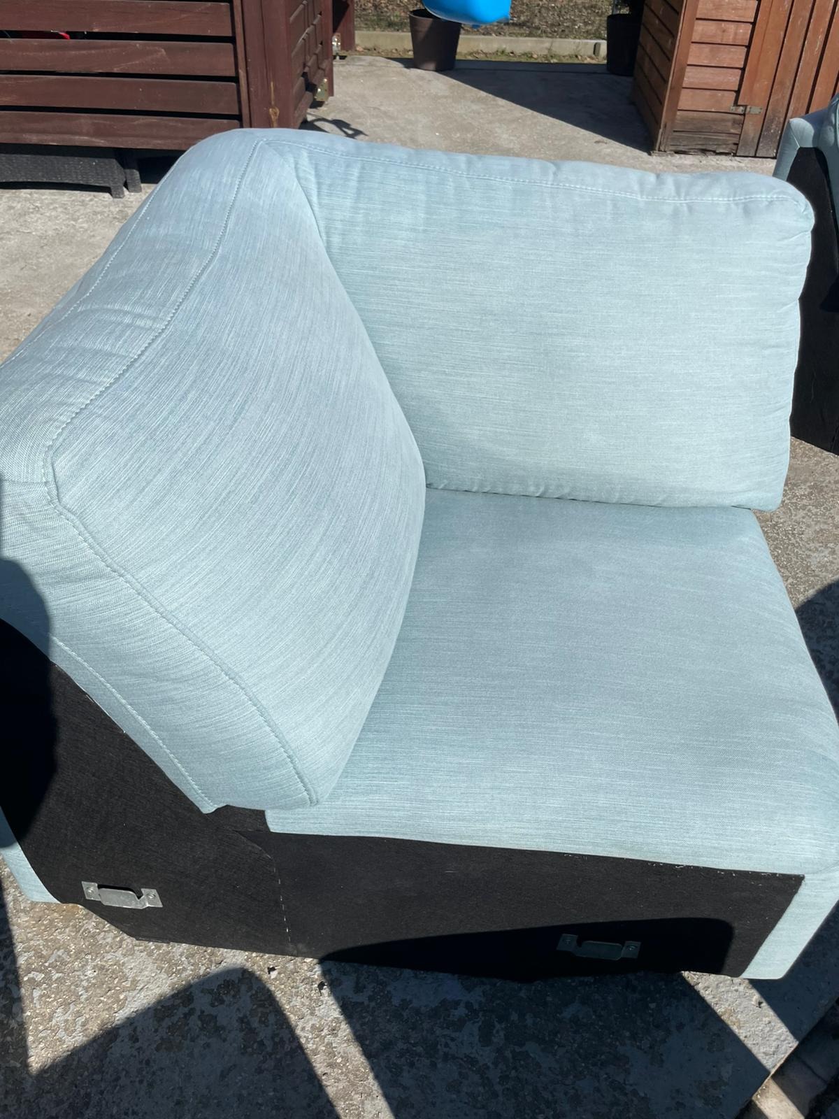 Jasnoniebieski narożnik z poduszką, widoczny na zewnątrz, z czarną podstawą i metalowymi uchwytami. Ujęcie z bliska, w dziennym świetle, ukazujące fakturę tkaniny.