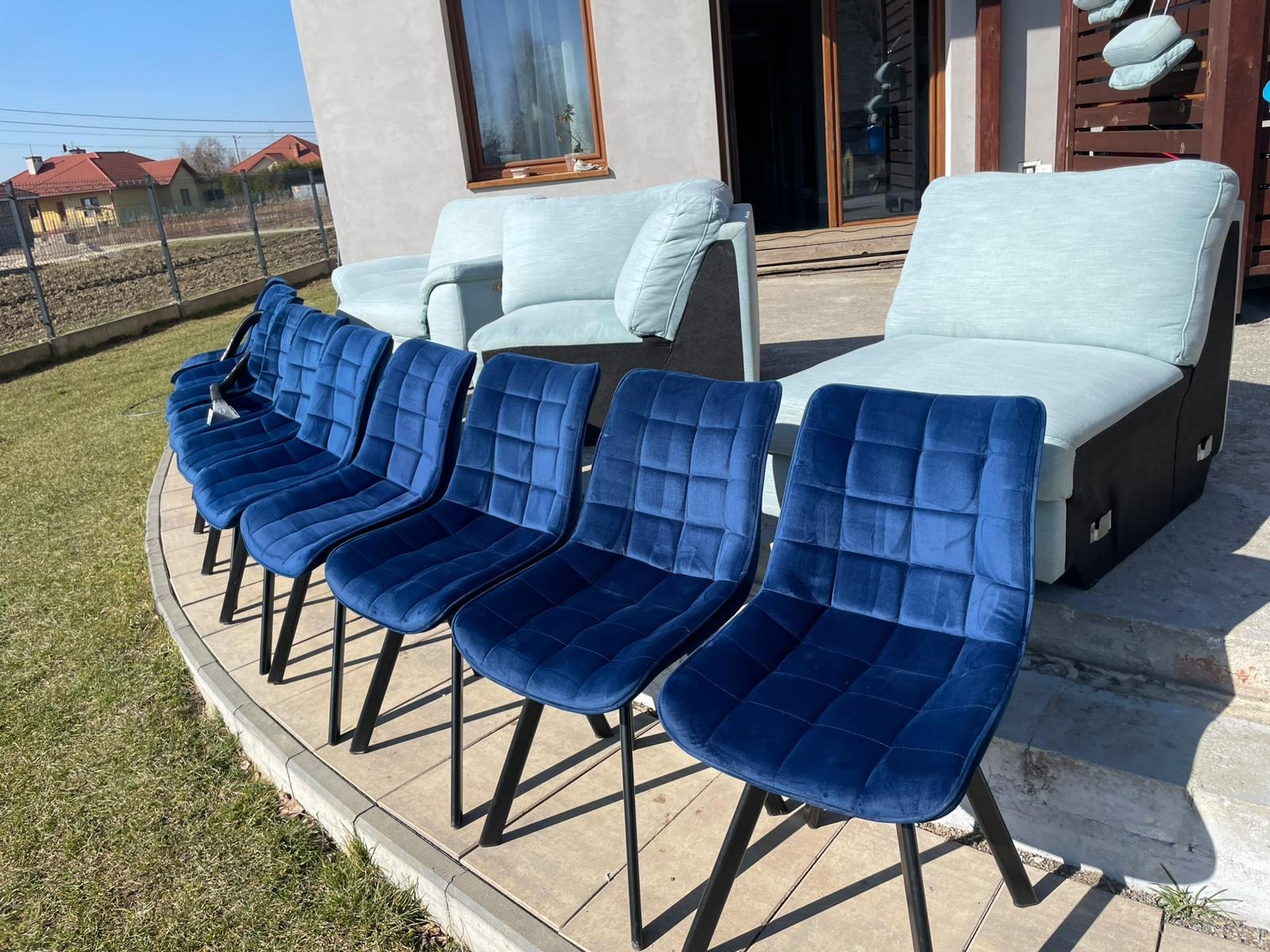 Rząd sześciu granatowych krzeseł z aksamitną tapicerką i czarnymi nogami na zewnątrz, z jasnoniebieską sofą i fotelem w tle, na tle domu.