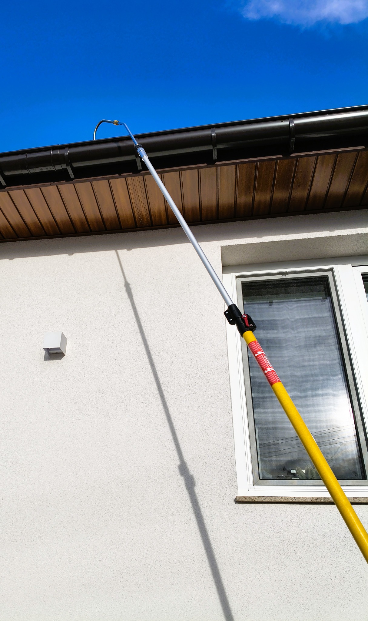 Czyszczenie rynny z poziomu gruntu w Łoniowie za pomocą teleskopowej lancy. Widoczna biała elewacja budynku, okno i brązowa podbitka dachowa.