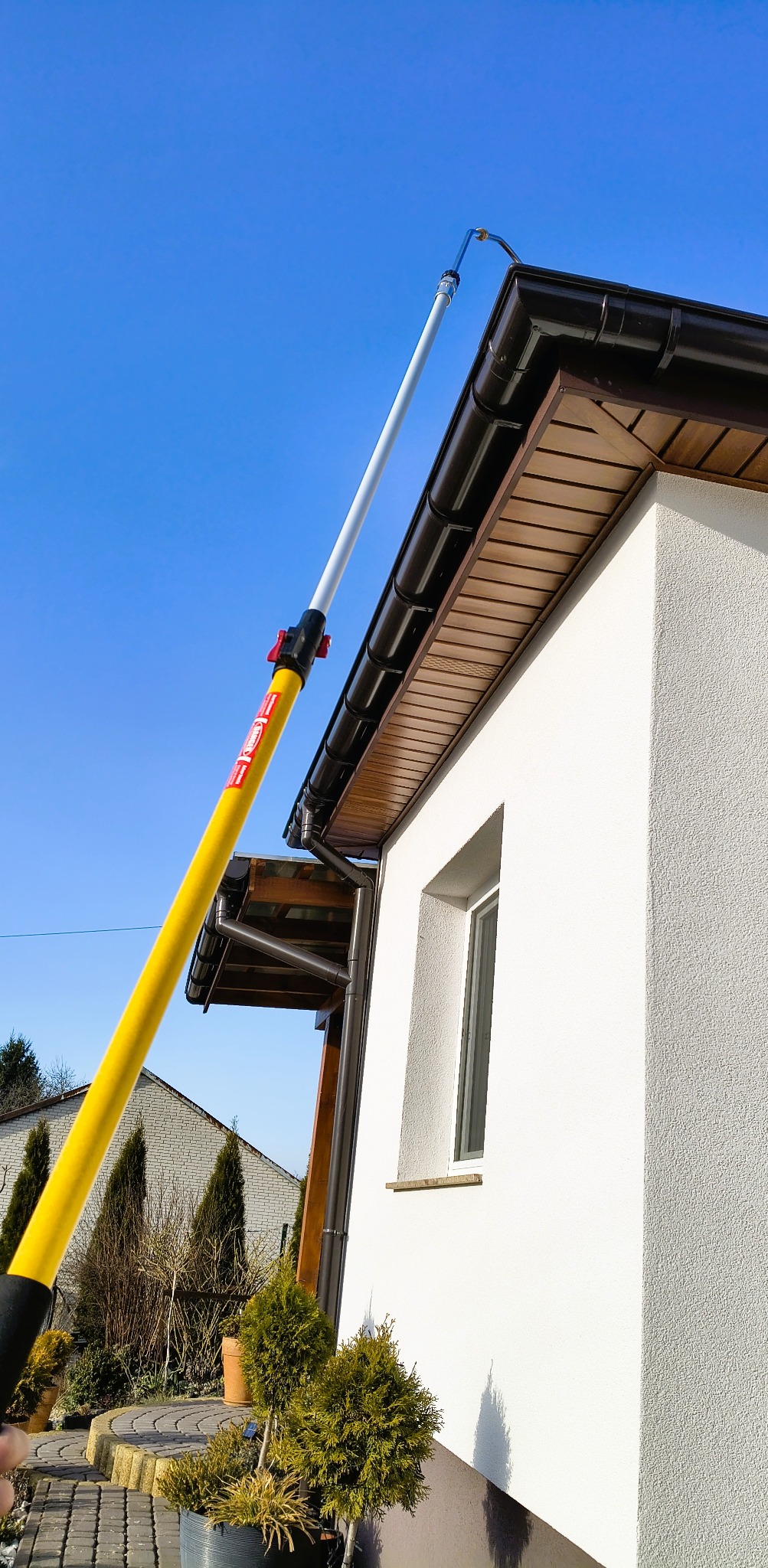 Czyszczenie rynny z ziemi za pomocą teleskopowego kija z dyszą. Biała elewacja domu, brązowe rynny i błękitne niebo w tle. Słoneczny dzień.