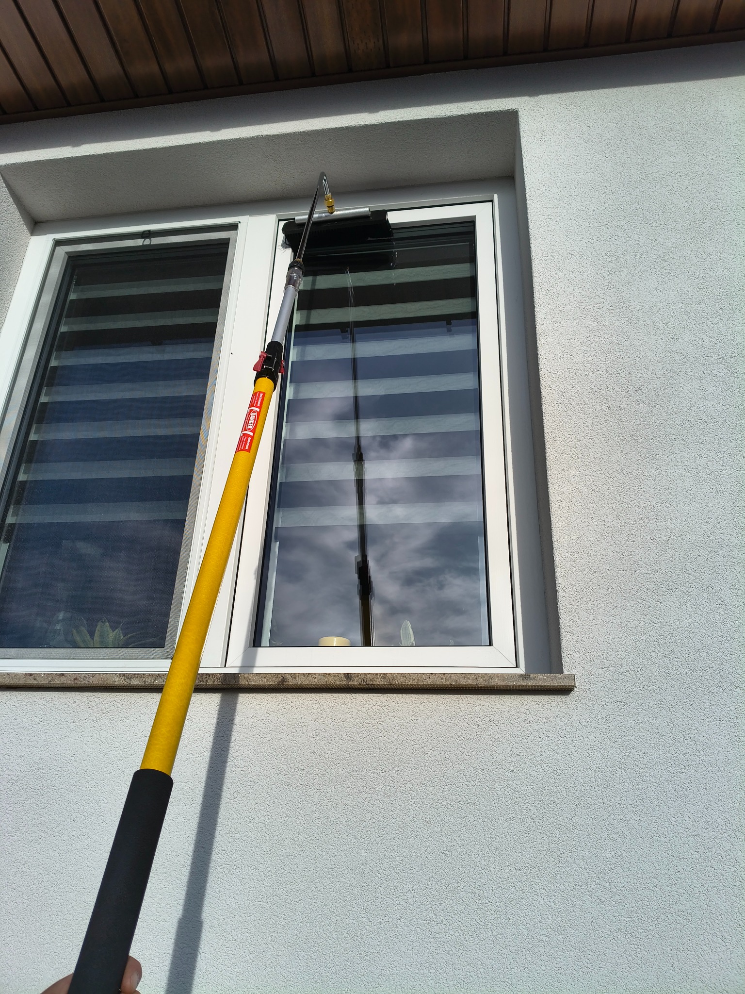 Białe okno z żaluzjami, czyszczone z zewnątrz żółtym kijem teleskopowym z czarną rączką. Widoczna faktura ściany budynku i odbicie nieba w szybie.