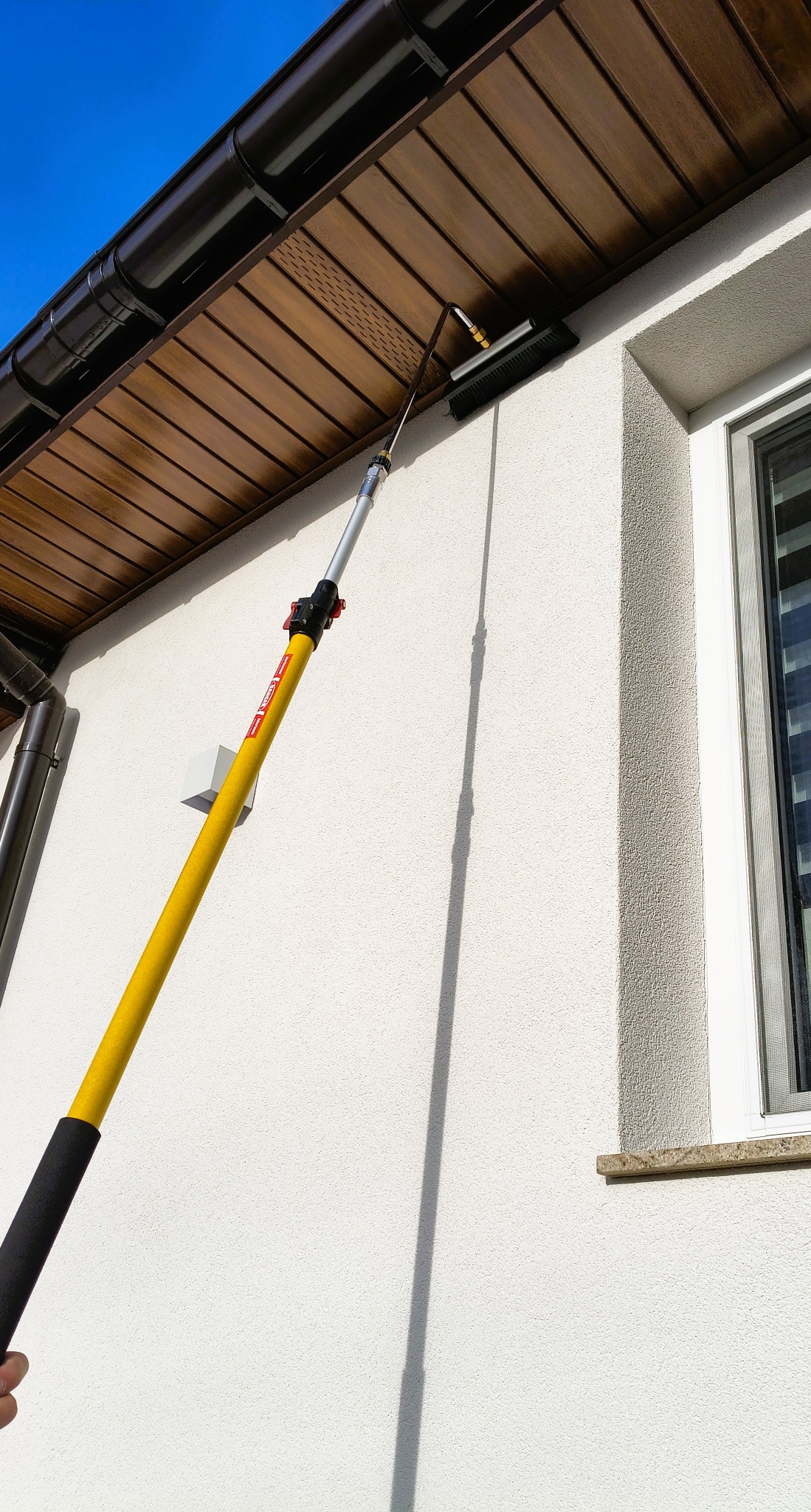Czyszczenie elewacji domu szczotką na teleskopowym kiju, widoczny fragment okna i podbitki dachowej. Ujęcie z dołu, słoneczny dzień.