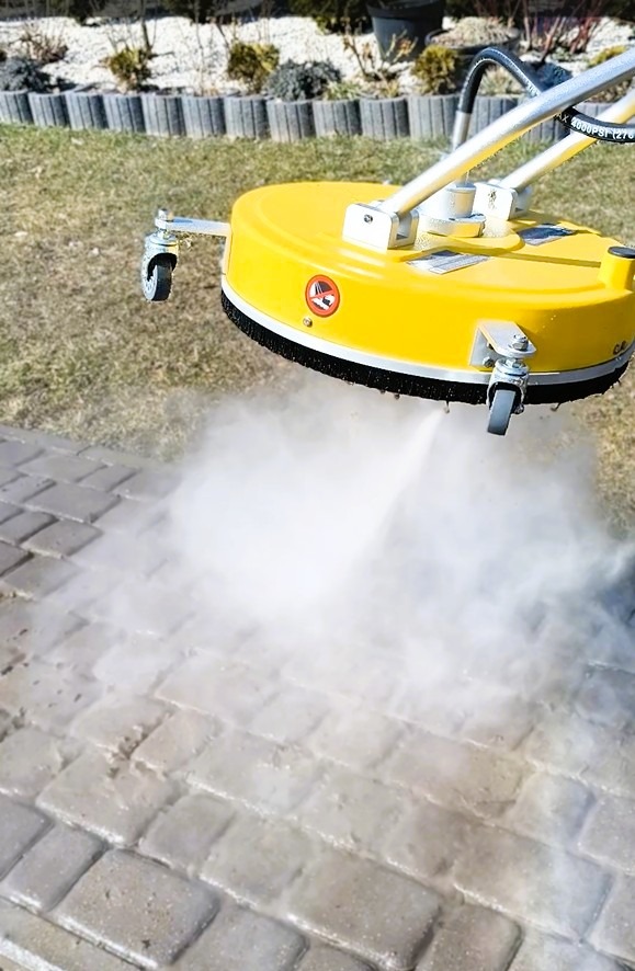 Żółta maszyna ciśnieniowa z dyszami rotacyjnymi czyści kostkę brukową na zewnątrz, tworząc obłok pary. Widoczne trawnik i rośliny w tle.