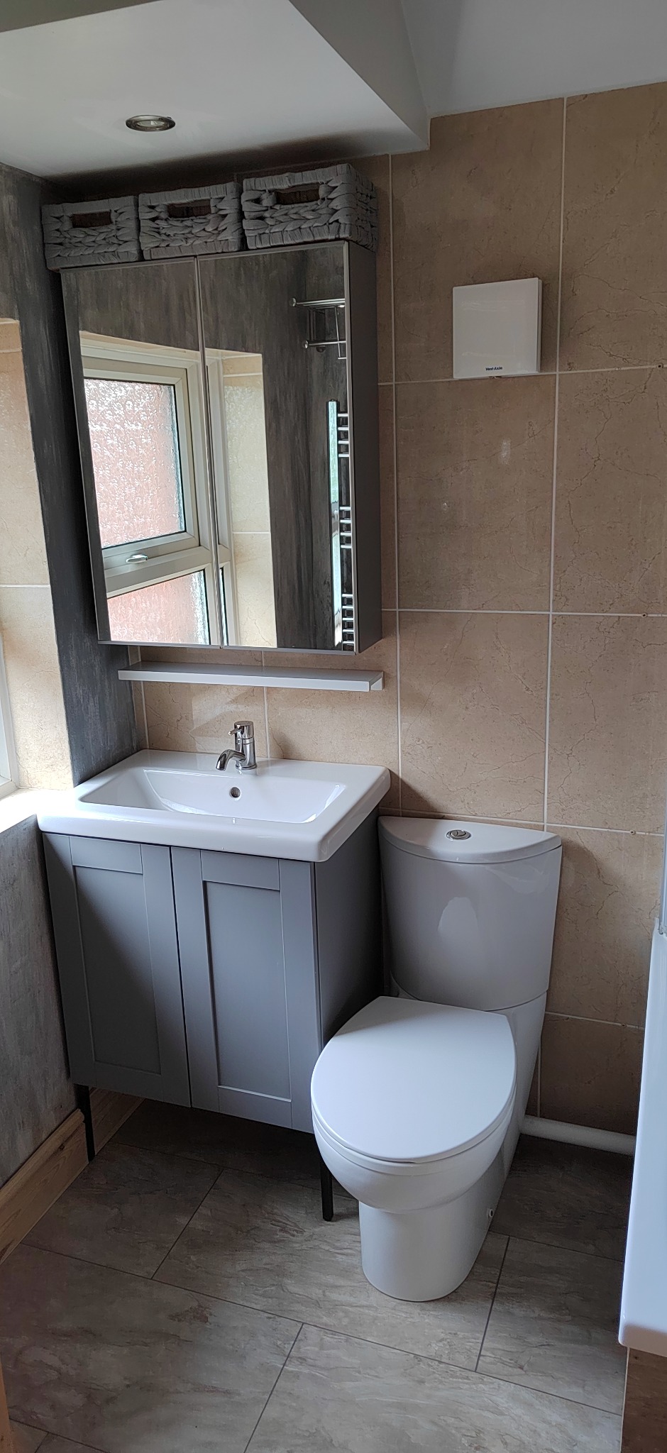 Jasna łazienka po remoncie: szara szafka z umywalką, toaleta, beżowe płytki, lustro z szafką i koszykami. Widok z przodu, neutralne oświetlenie.