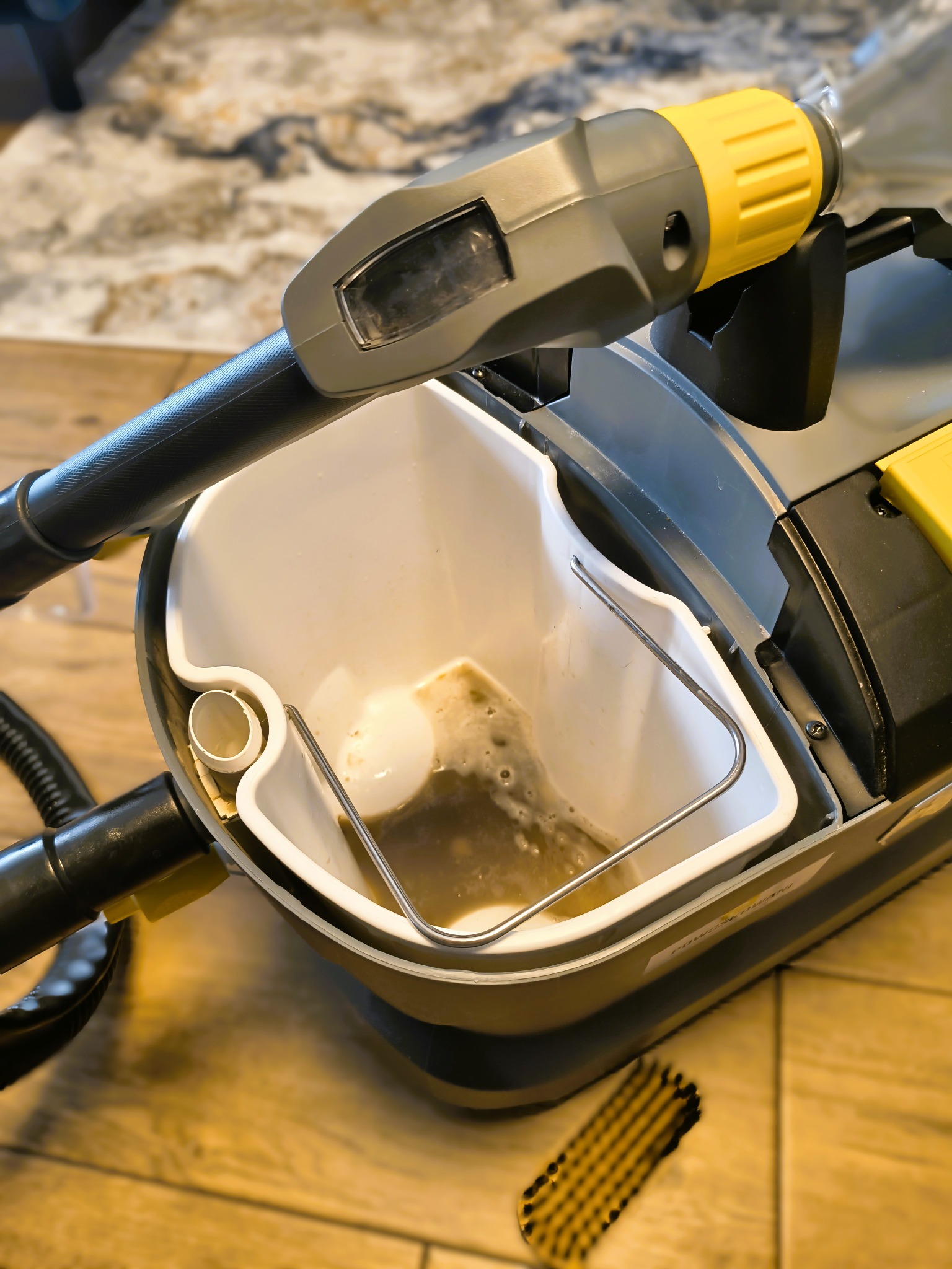 Zbliżenie na zbiornik odkurzacza piorącego z brudną wodą po czyszczeniu, obok szczotka. Widoczna struktura podłogi i fragment dywanu w tle. Sprzęt do ekstrakcyjnego czyszczenia.