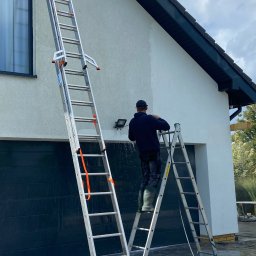 MK Błysk - Pracownik na drabinie myje elewację domu z garażem, używając myjki ciśnieniowej. Widoczne zacieki i woda spływająca po ścianie. Ujęcie z dołu.