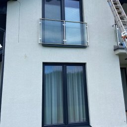 MK Błysk - Jasna elewacja domu z dwoma oknami i balkonem z szklaną balustradą. Przy ścianie oparta drabina. Widoczny system alarmowy.
