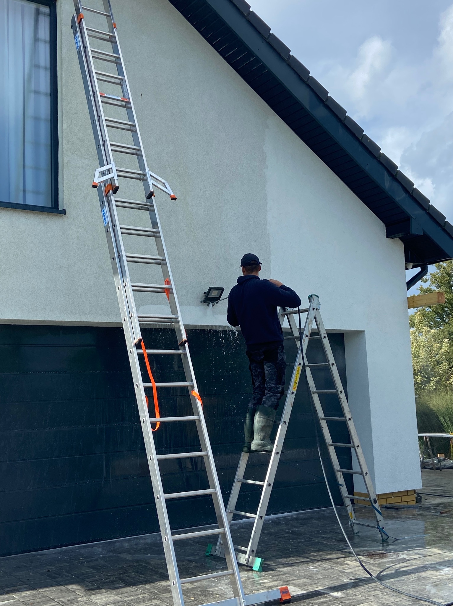 Pracownik na drabinie myje elewację domu z garażem, używając myjki ciśnieniowej. Widoczne zacieki i woda spływająca po ścianie. Ujęcie z dołu.