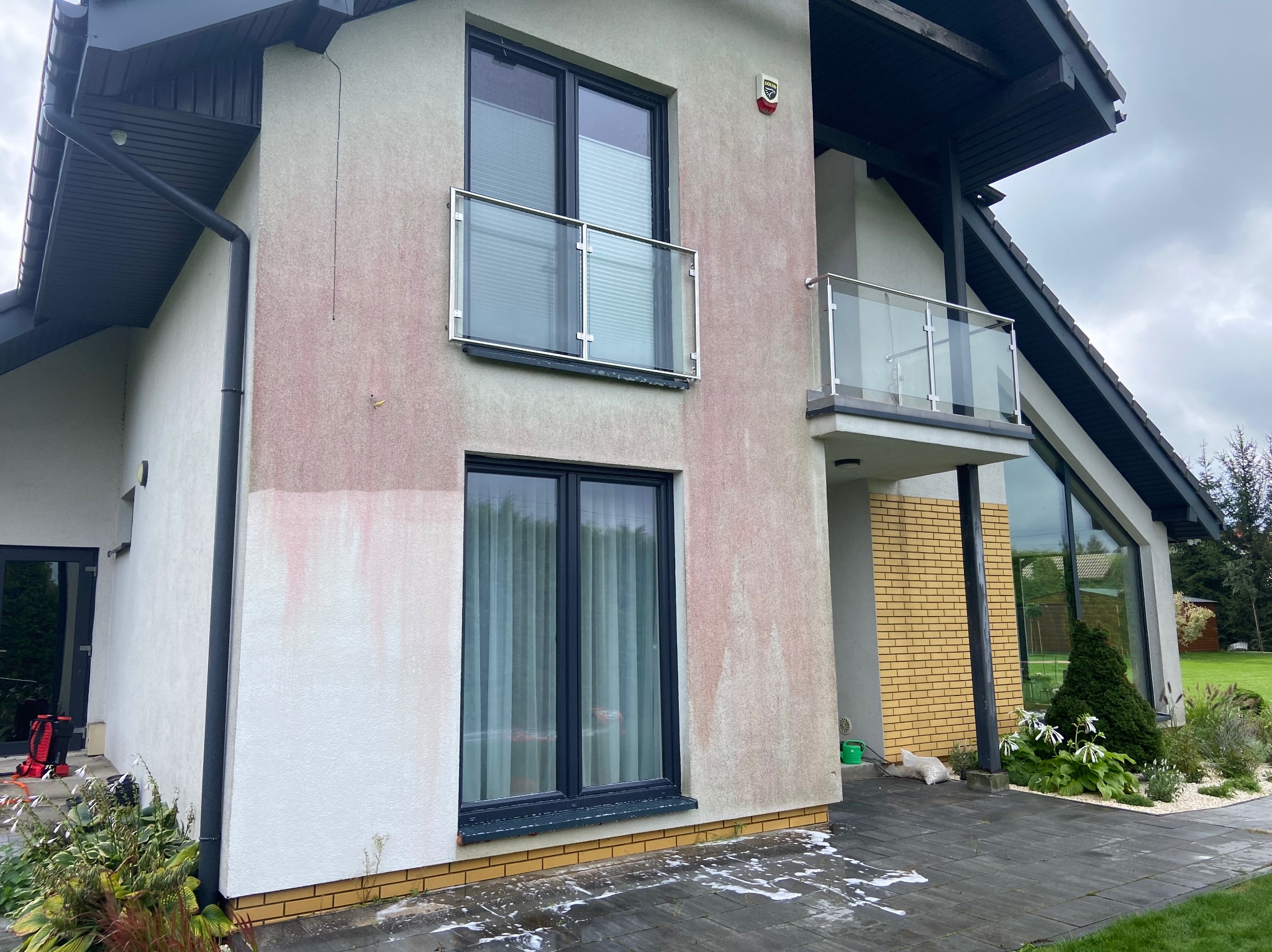 Dwupiętrowy dom z elewacją w trakcie czyszczenia. Widoczne zacieki i pozostałości po myciu na jasnej fasadzie. Nowoczesna architektura z balkonami i dużymi oknami.