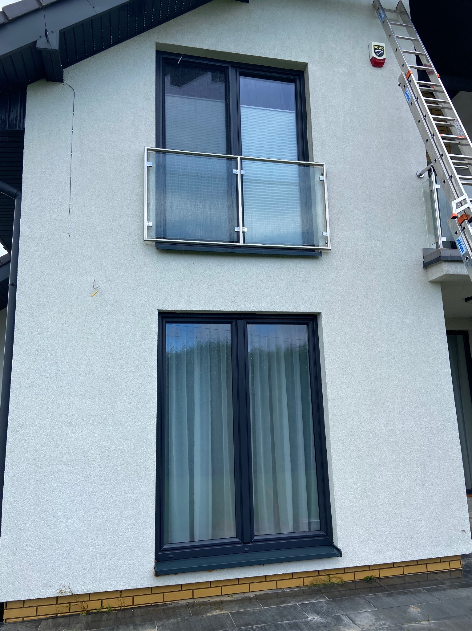 Jasna elewacja domu z dwoma oknami i balkonem z szklaną balustradą. Przy ścianie oparta drabina. Widoczny system alarmowy.