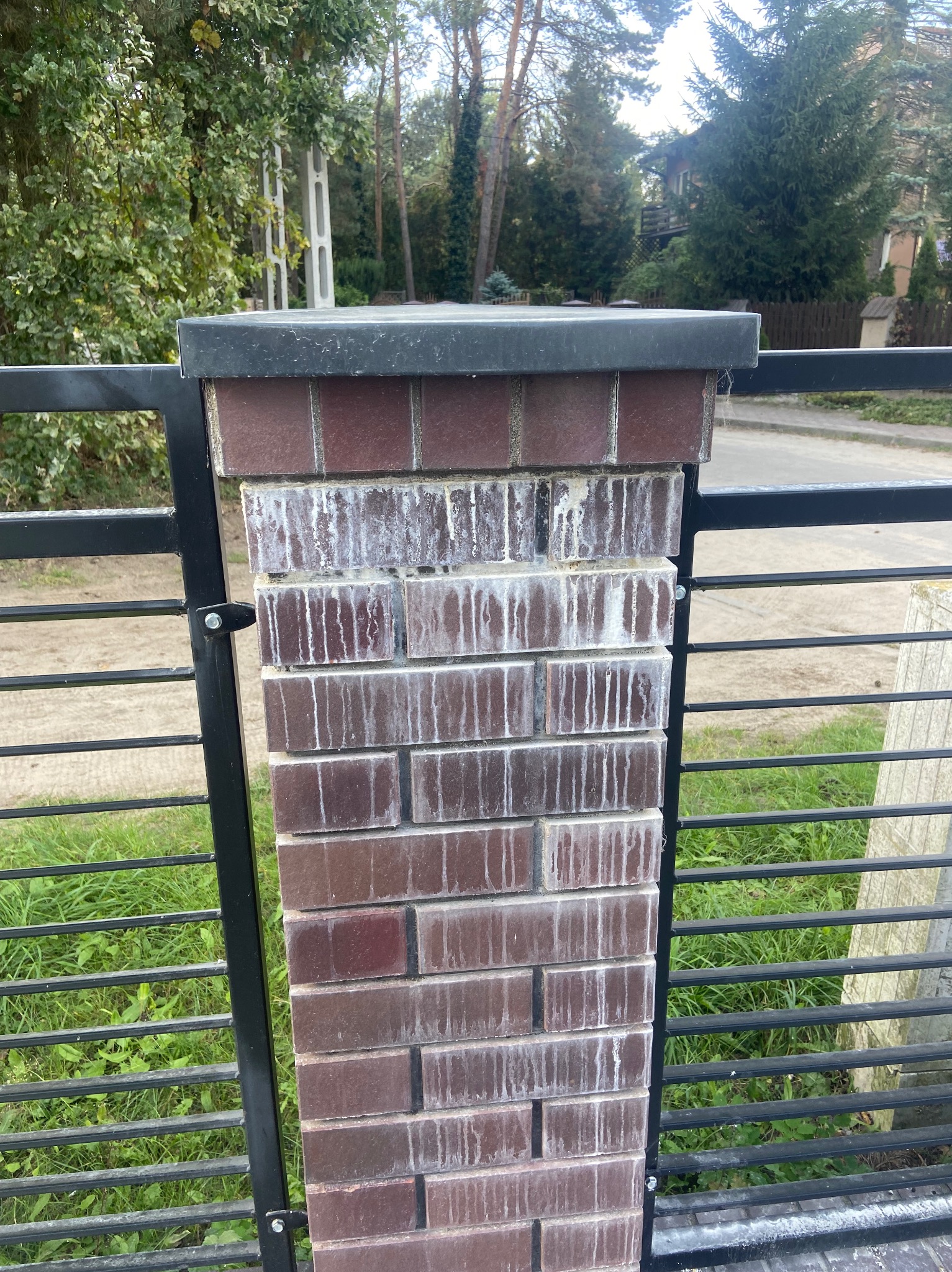 Fragment ogrodzenia z ceglanym słupkiem z widocznymi zaciekami, czarna metalowa konstrukcja, zieleń w tle. Słupek z cegły klinkierowej.