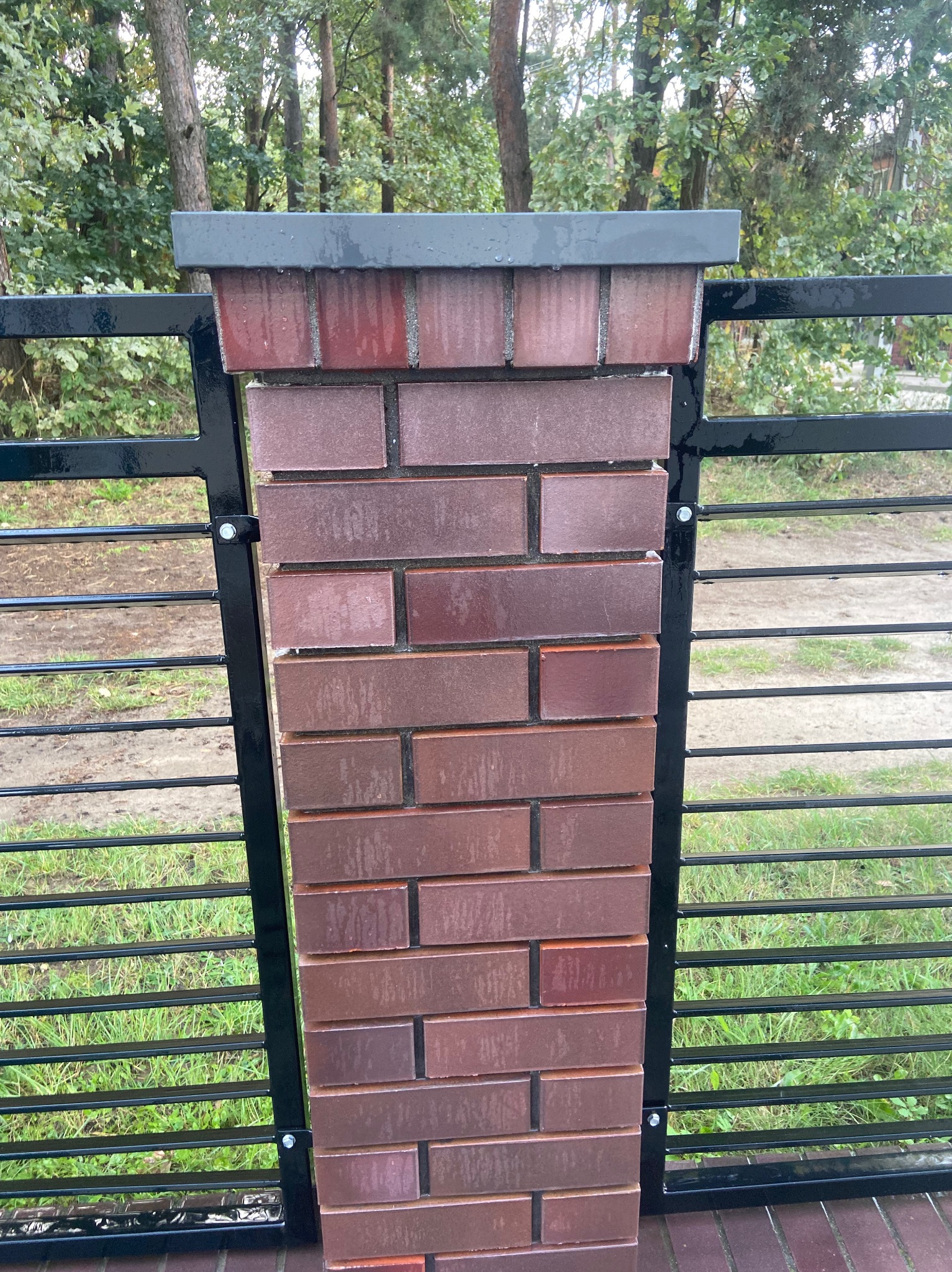 Ceglany słupek ogrodzeniowy z czarnym metalowym wypełnieniem, widok z bliska na tle zieleni. Słupek zwieńczony szarym daszkiem, całość wygląda na świeżo po deszczu.