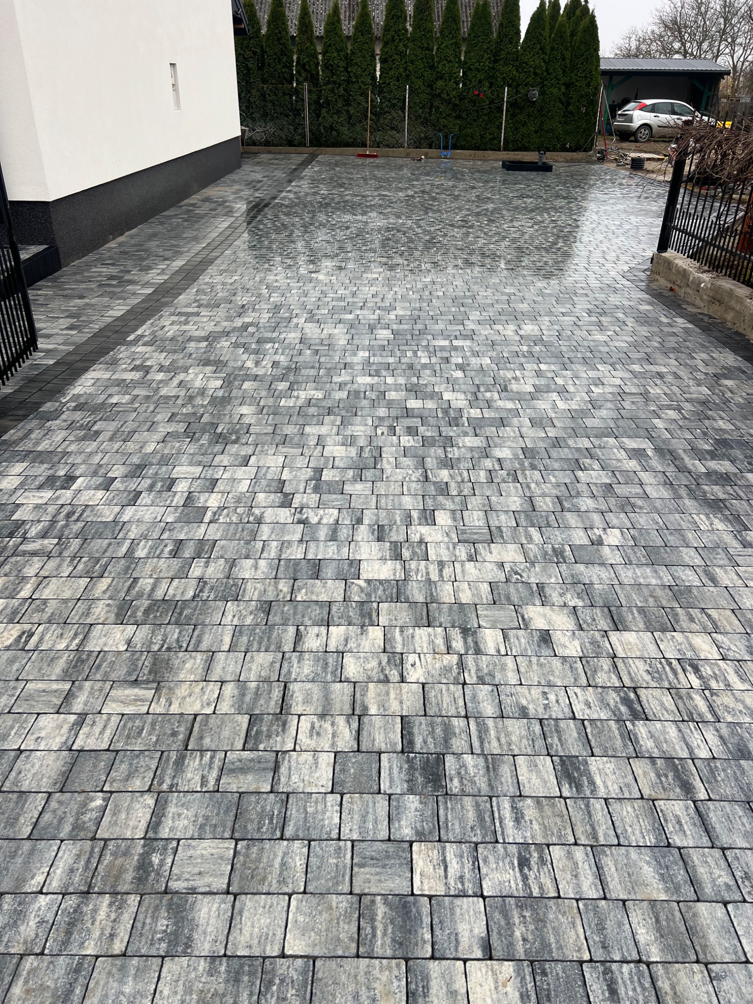 Ułożona, szara kostka brukowa na podjeździe, z widocznym fragmentem budynku i zieleni w tle. Kostka ułożona w regularny wzór, lekko mokra, odbija światło.