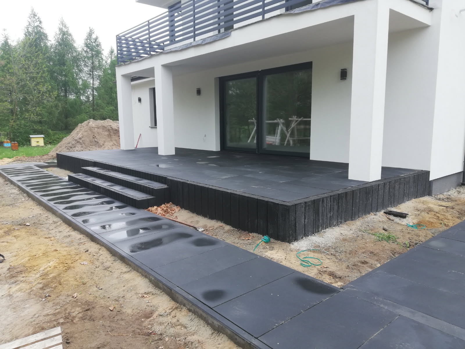 Nowoczesny taras z ciemnej kostki brukowej, schody i ścieżka prowadząca wzdłuż budynku. Widoczne detale wykończenia i otoczenie w trakcie prac.