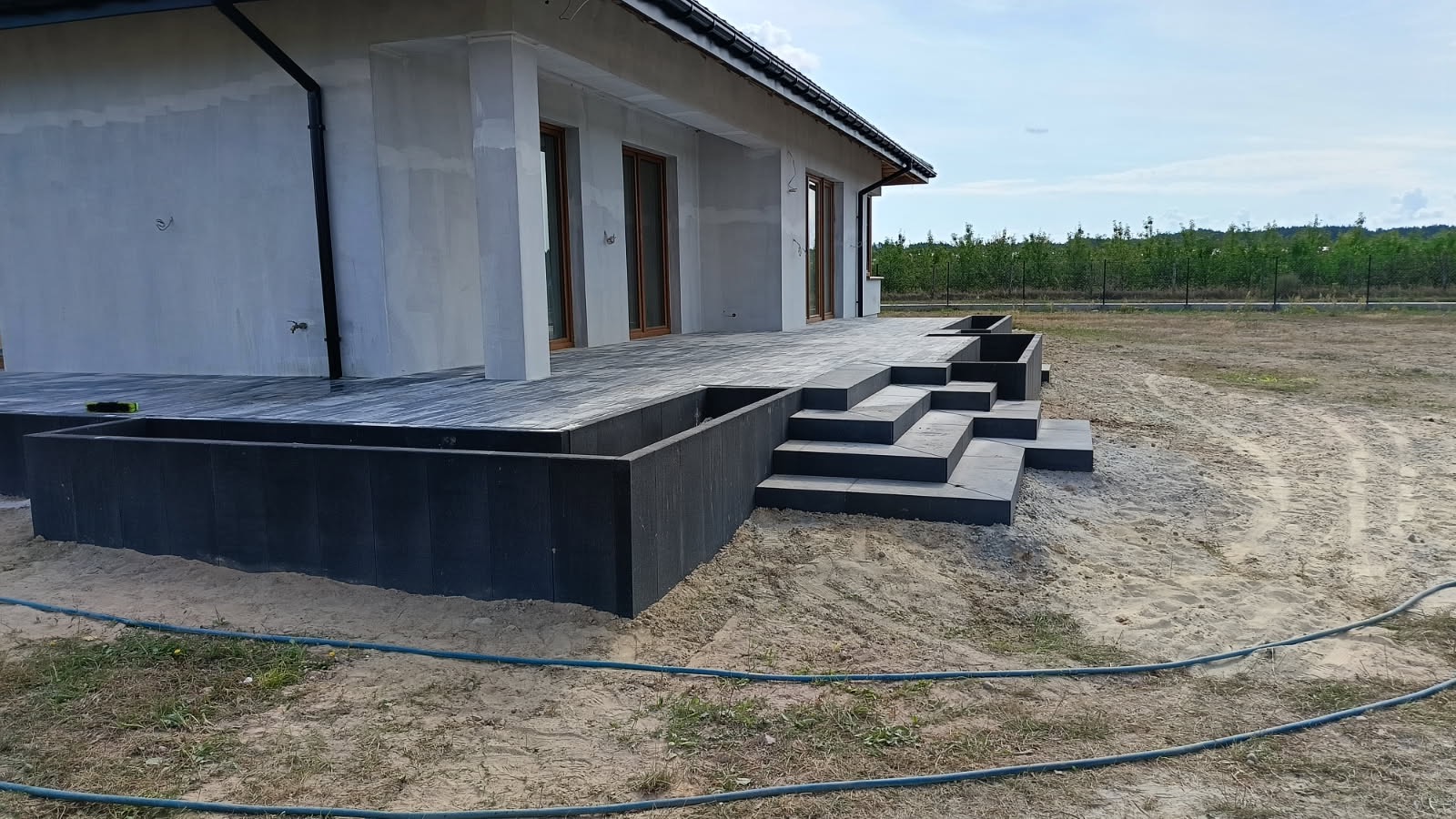 Nowoczesny taras z kostki brukowej w odcieniach szarości, z niskim murkiem i schodami, przylegający do surowego budynku w trakcie wykończenia.