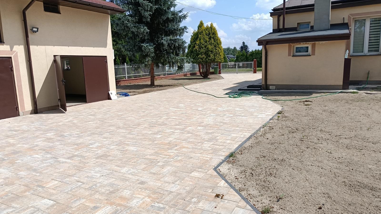 Beżowa kostka brukowa ułożona na podjeździe między budynkami, z widocznym wężem ogrodowym i fragmentem niezagospodarowanego terenu. Jasne, słoneczne oświetlenie.