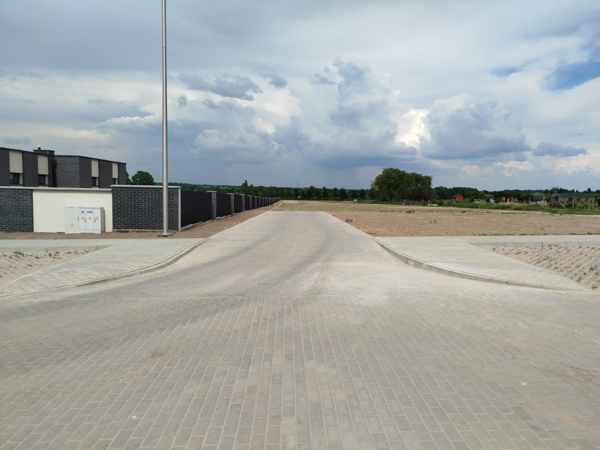 Nowa, szara kostka brukowa tworząca drogę dojazdową, z widocznymi obrzeżami i elementami krajobrazu w tle, pod pochmurnym niebem.