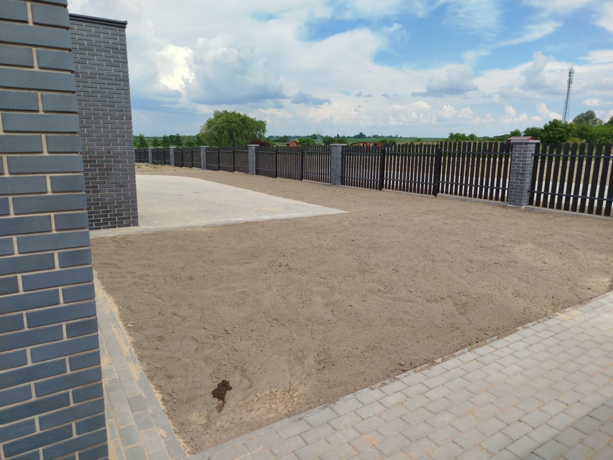 Przygotowany teren pod trawnik z obrzeżami z kostki i ogrodzeniem z ceglanymi słupkami. Widok na krajobraz zza ogrodzenia.